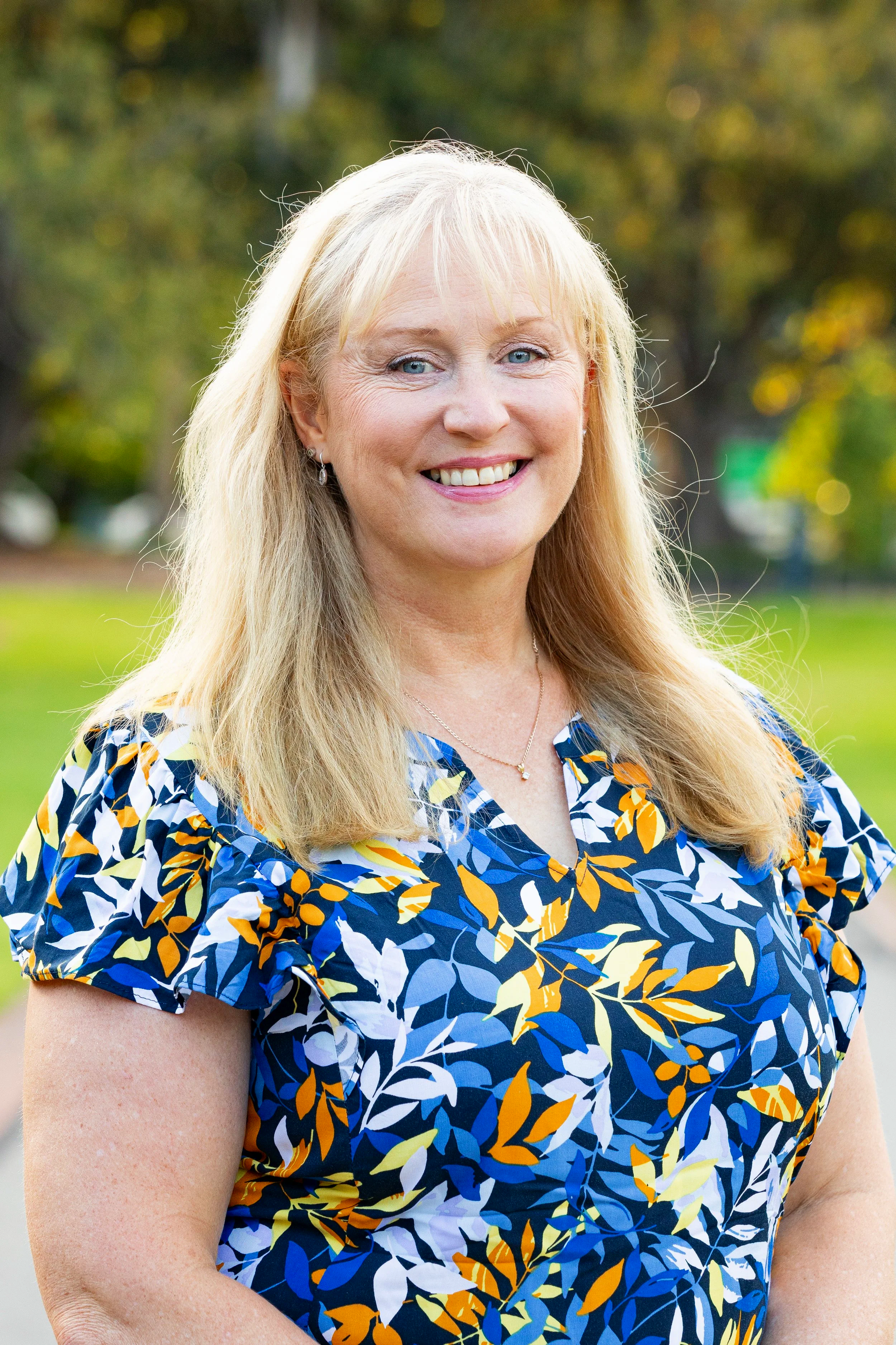 A middle-aged woman with blonde hair and blue eyes, smiling, outdoors in a park with trees in the background, wearing a colorful floral blouse.