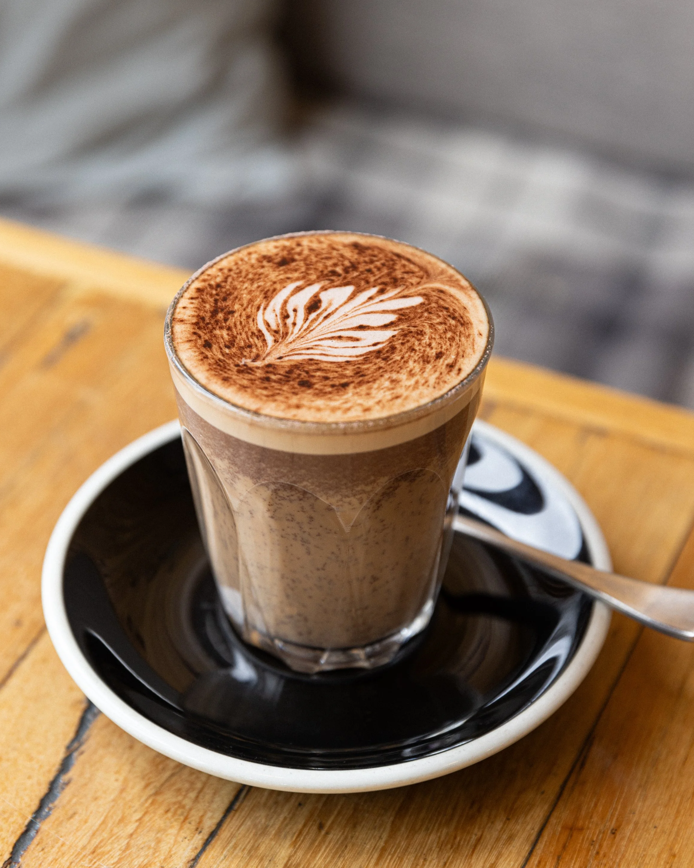 A glass of caramel macchiato with latte art on top, placed on a black saucer with a spoon, on a wooden table.