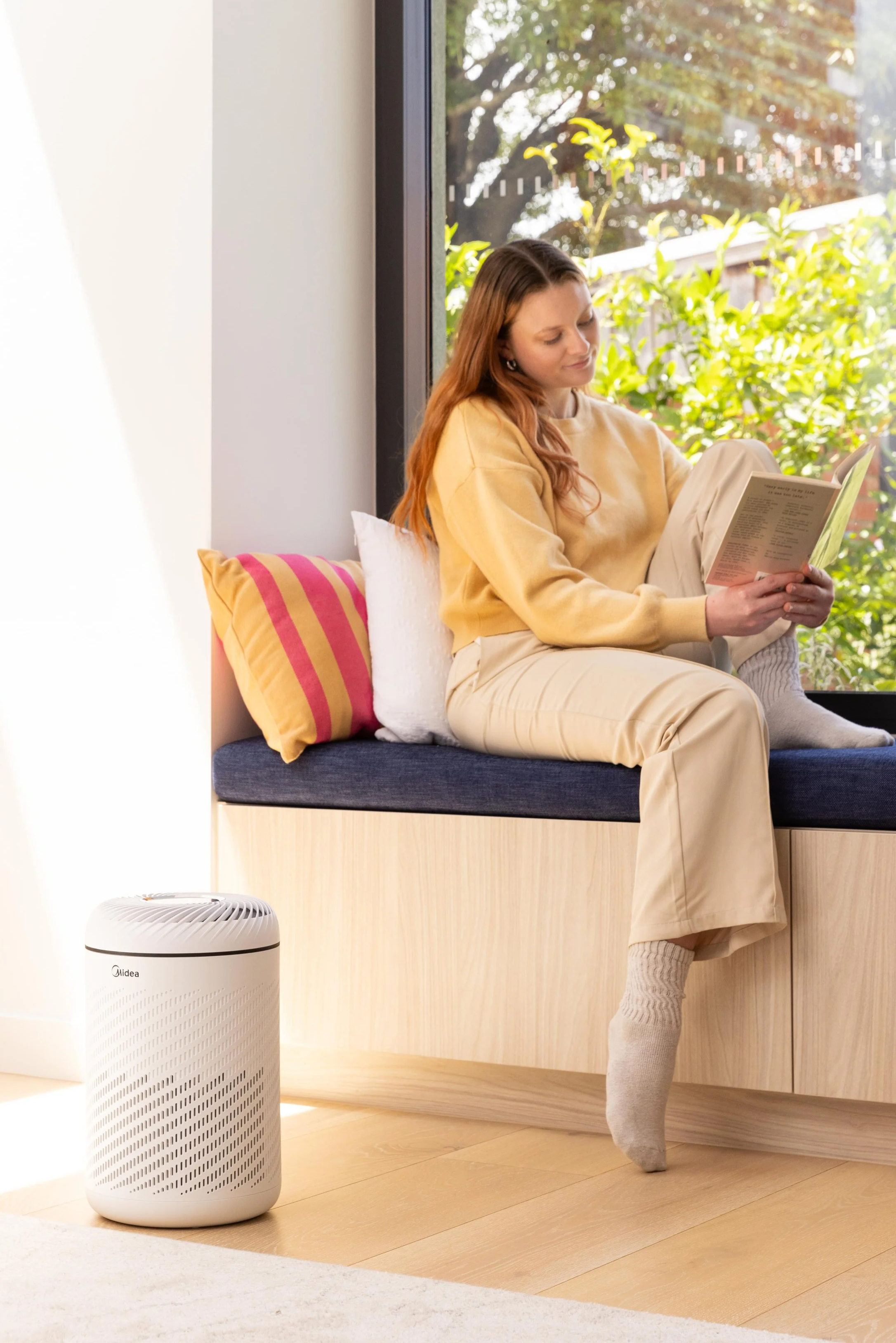 A woman sitting on a window seat reading a book, with a large window and outdoor greenery behind her. A white air purifier is on the floor nearby.