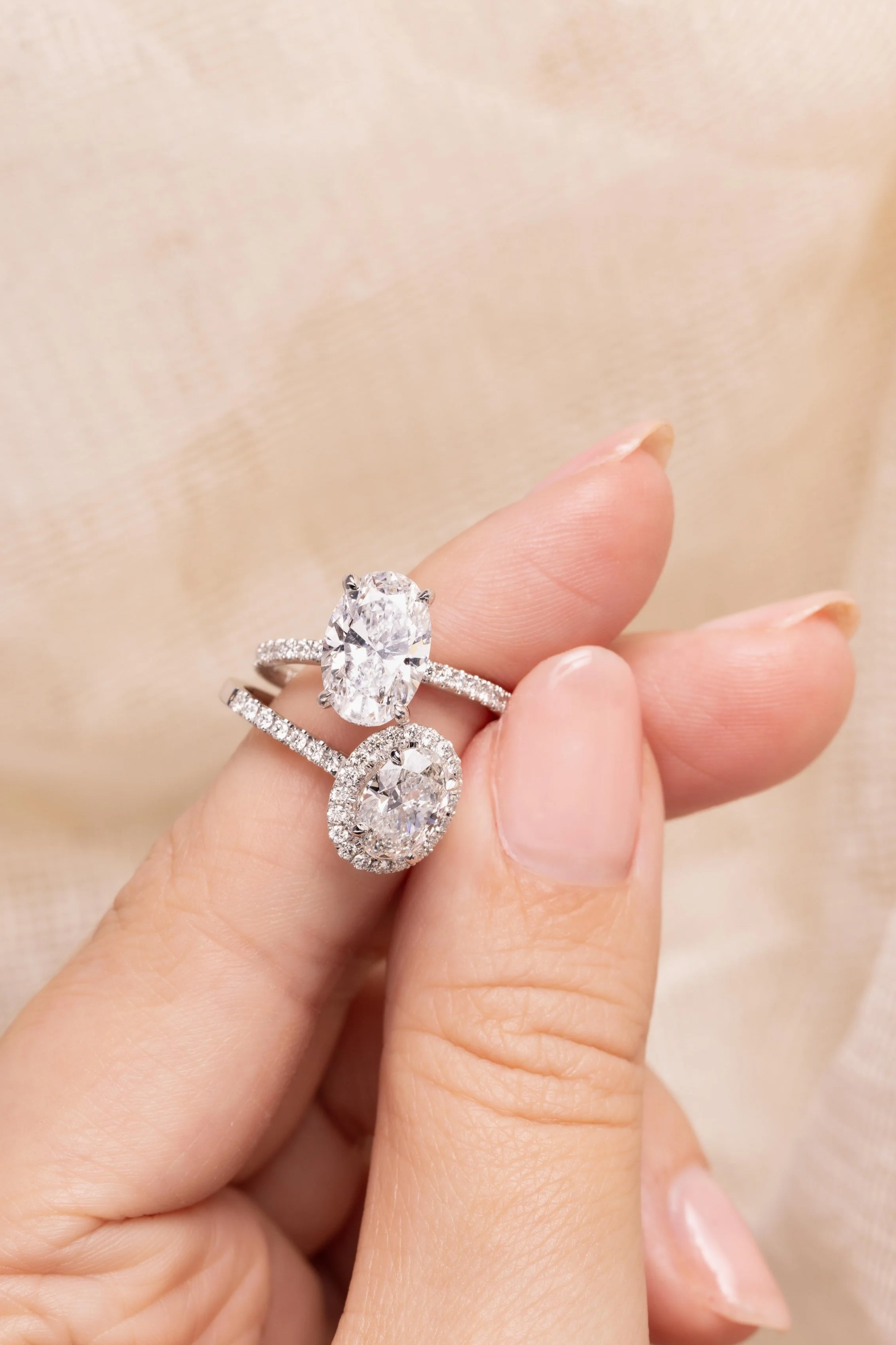 Person holding two diamond rings with oval and round cut diamonds, set in silver bands, against a beige fabric background.