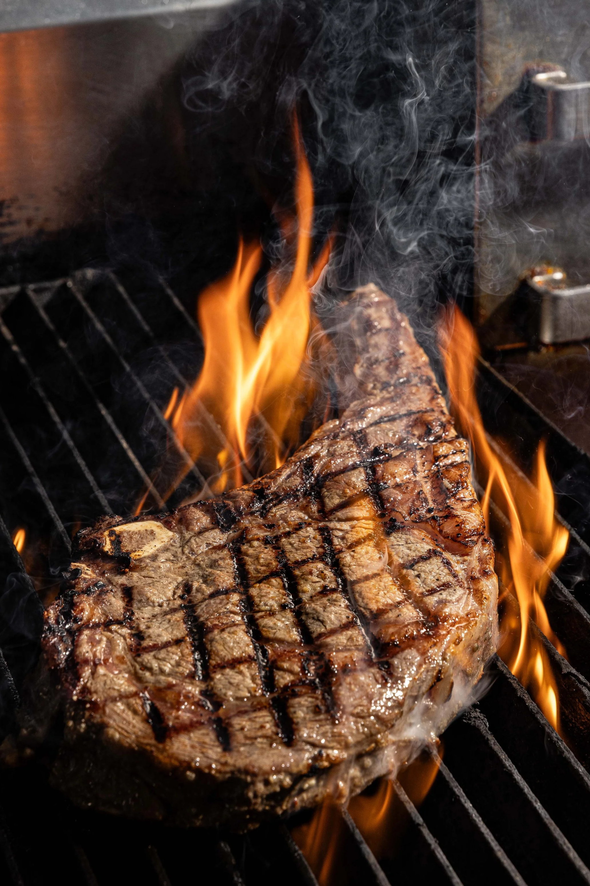 A grilled steak cooking on an outdoor grill with flames and smoke.