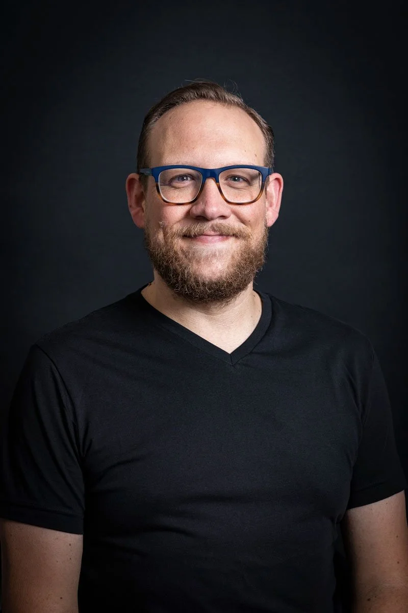 A man with glasses, a beard, and short hair posing against a dark background.
