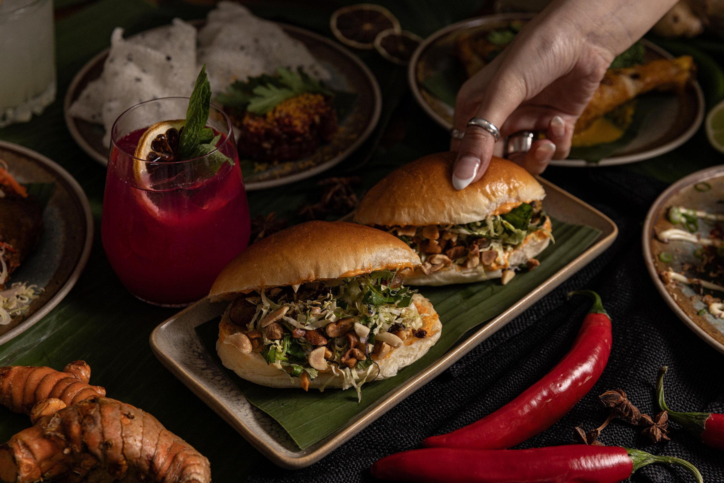 A table with two sandwiches with leafy greens and nuts, a red chili pepper, a glass of colorful beverage garnished with a lemon slice, and various plates of food in the background.
