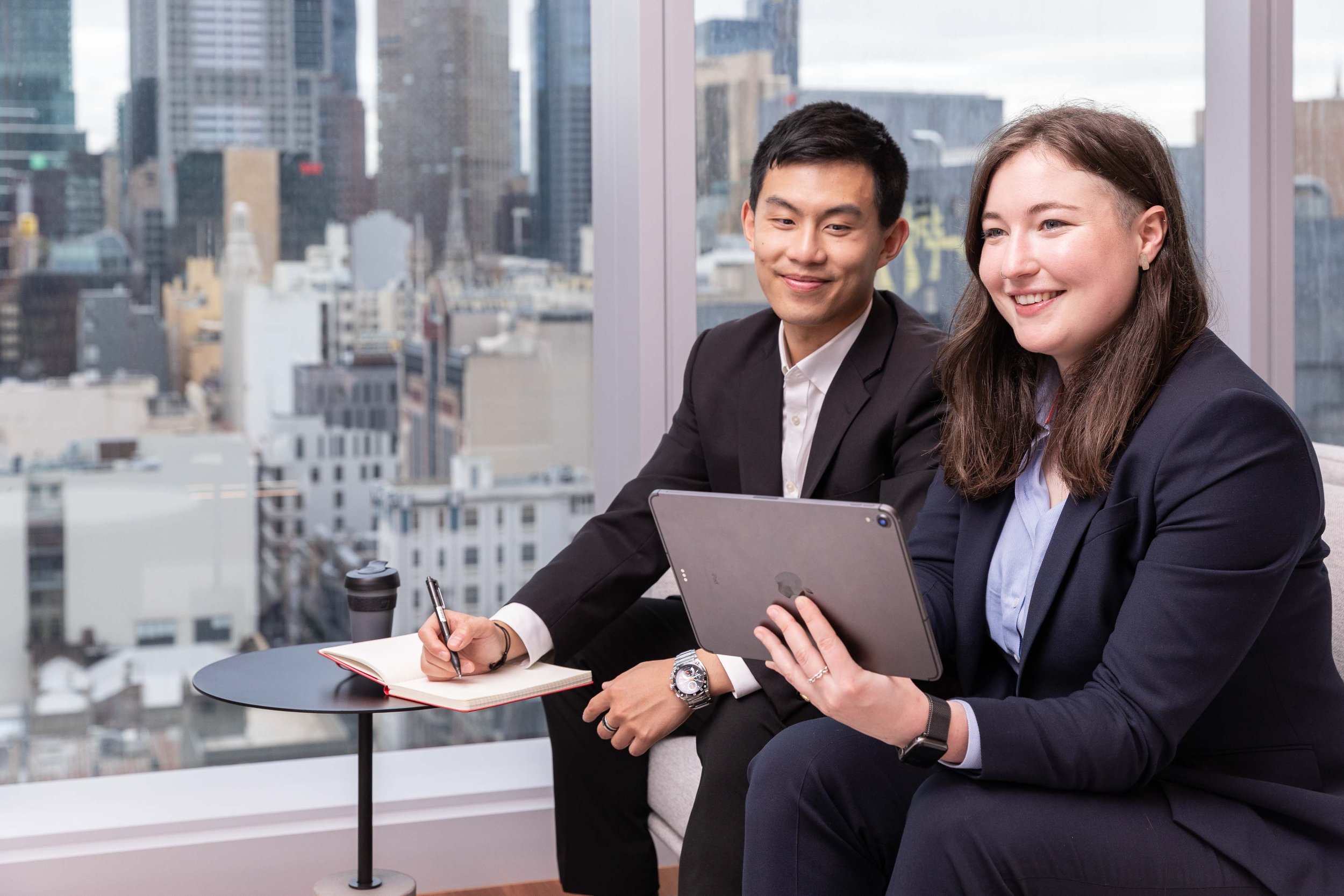 Two business professionals, a man and a woman, sitting in a modern office with a city skyline in the background, engaged in a discussion with electronic devices and notes.