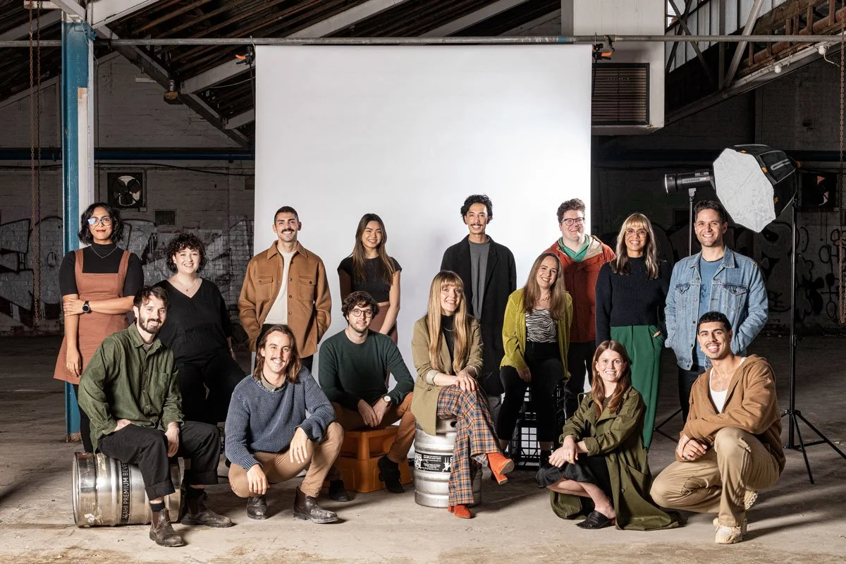 A diverse group of 15 people posing for a photo in a photography studio with a white backdrop, professional lighting equipment, and industrial decor, all smiling and dressed casually.