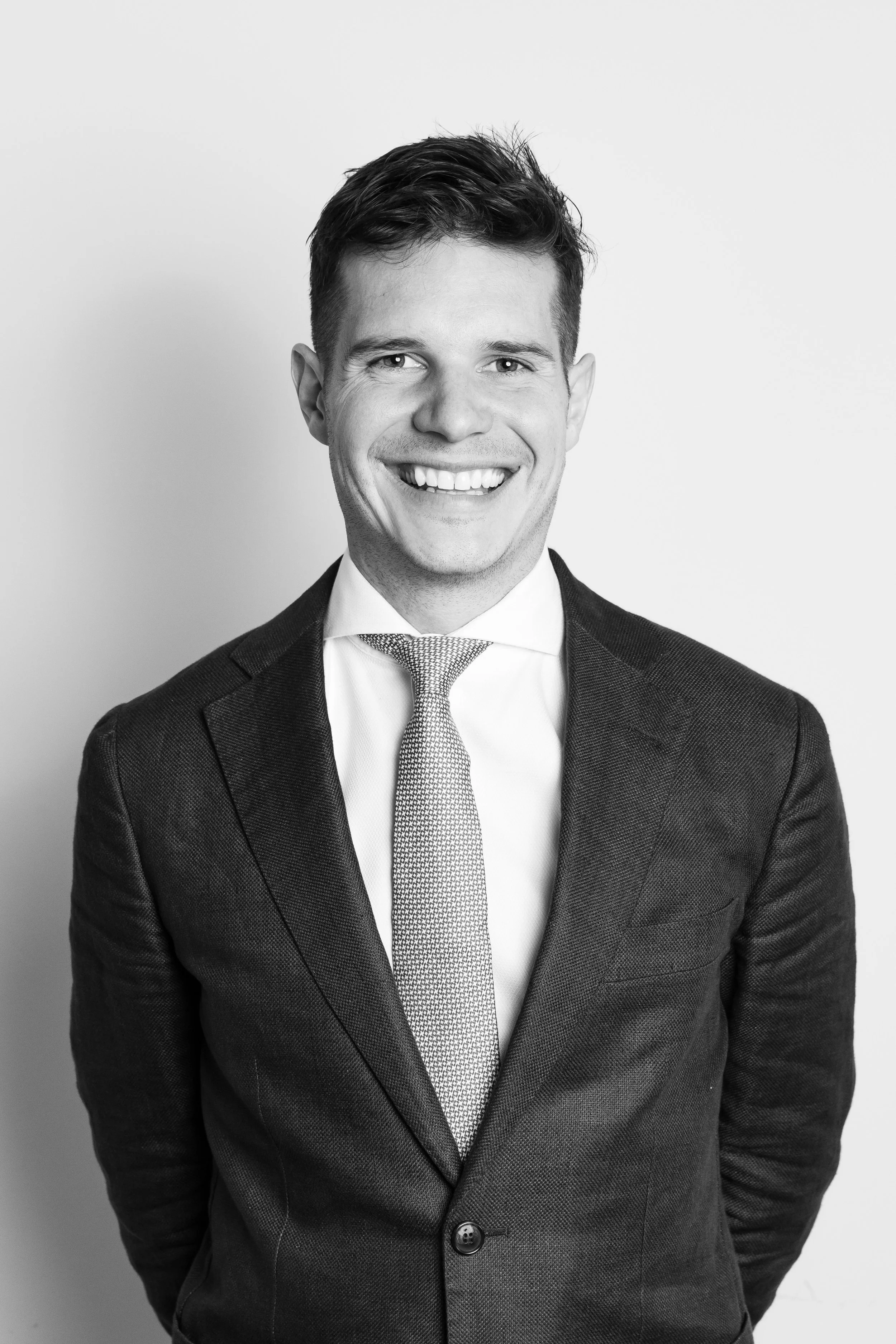 Black and white professional headshot of a smiling young man in a suit and tie, standing against a plain background.