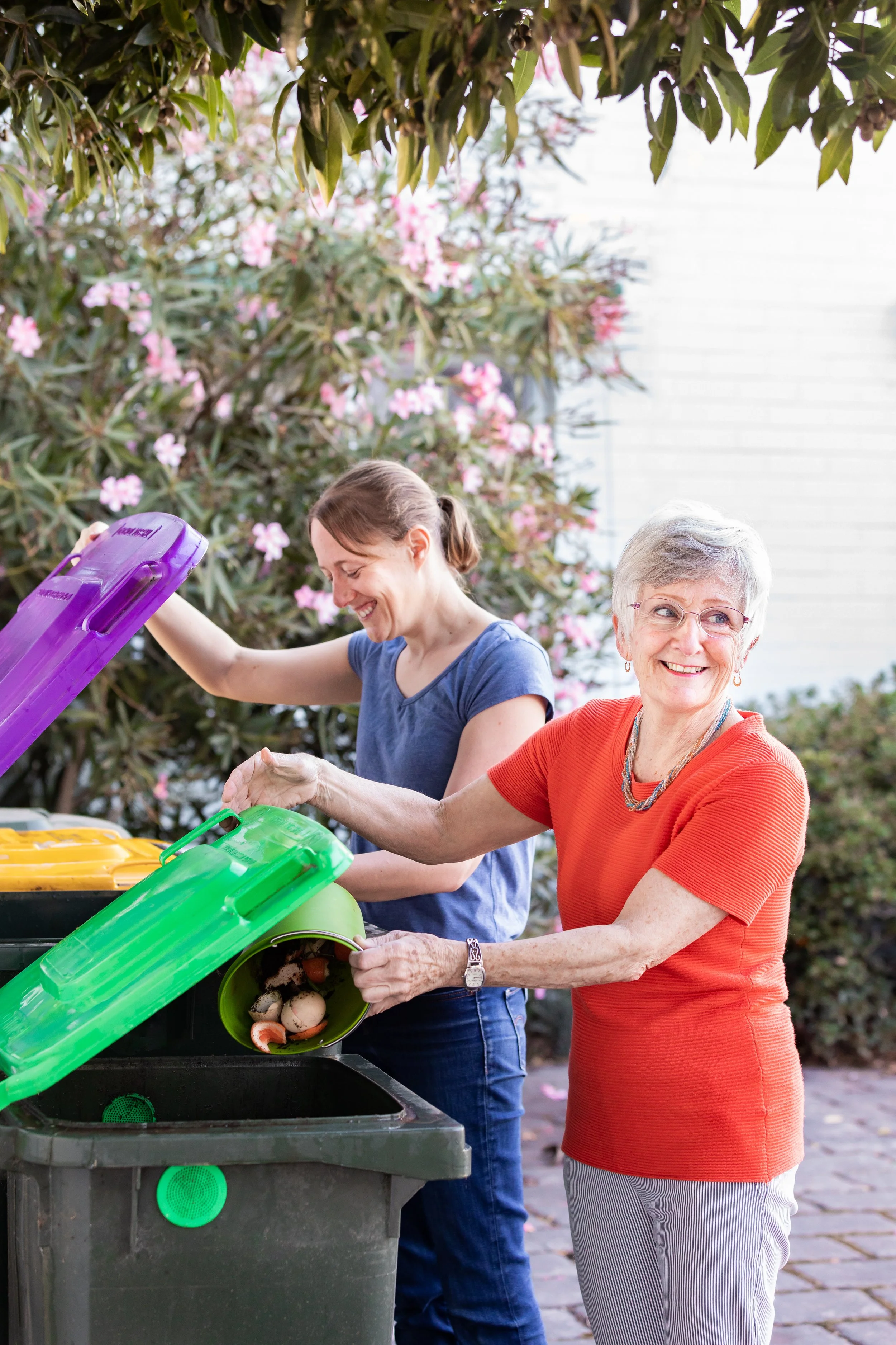 A woman and an elderly woman are sorting trash into recycling bins outdoors, with blooming pink flowers and shrubs in the background.
