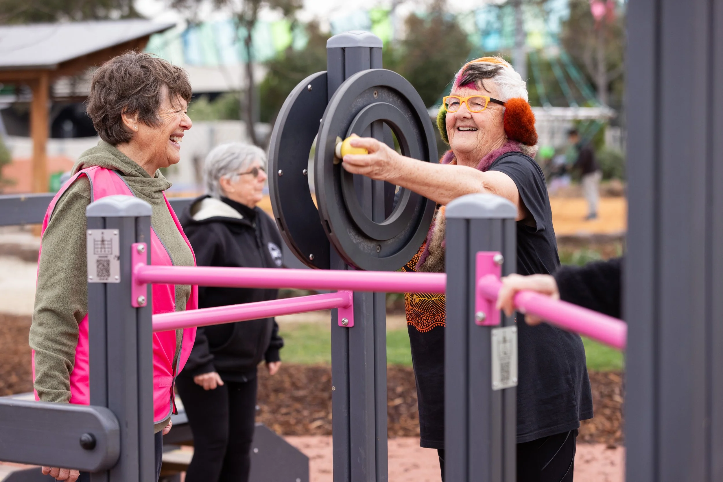 Two elderly women smiling and laughing at an outdoor park fitness equipment. One woman with colorful earmuffs and eyeglasses is reaching into a circular opening, while another woman in a pink vest looks on.