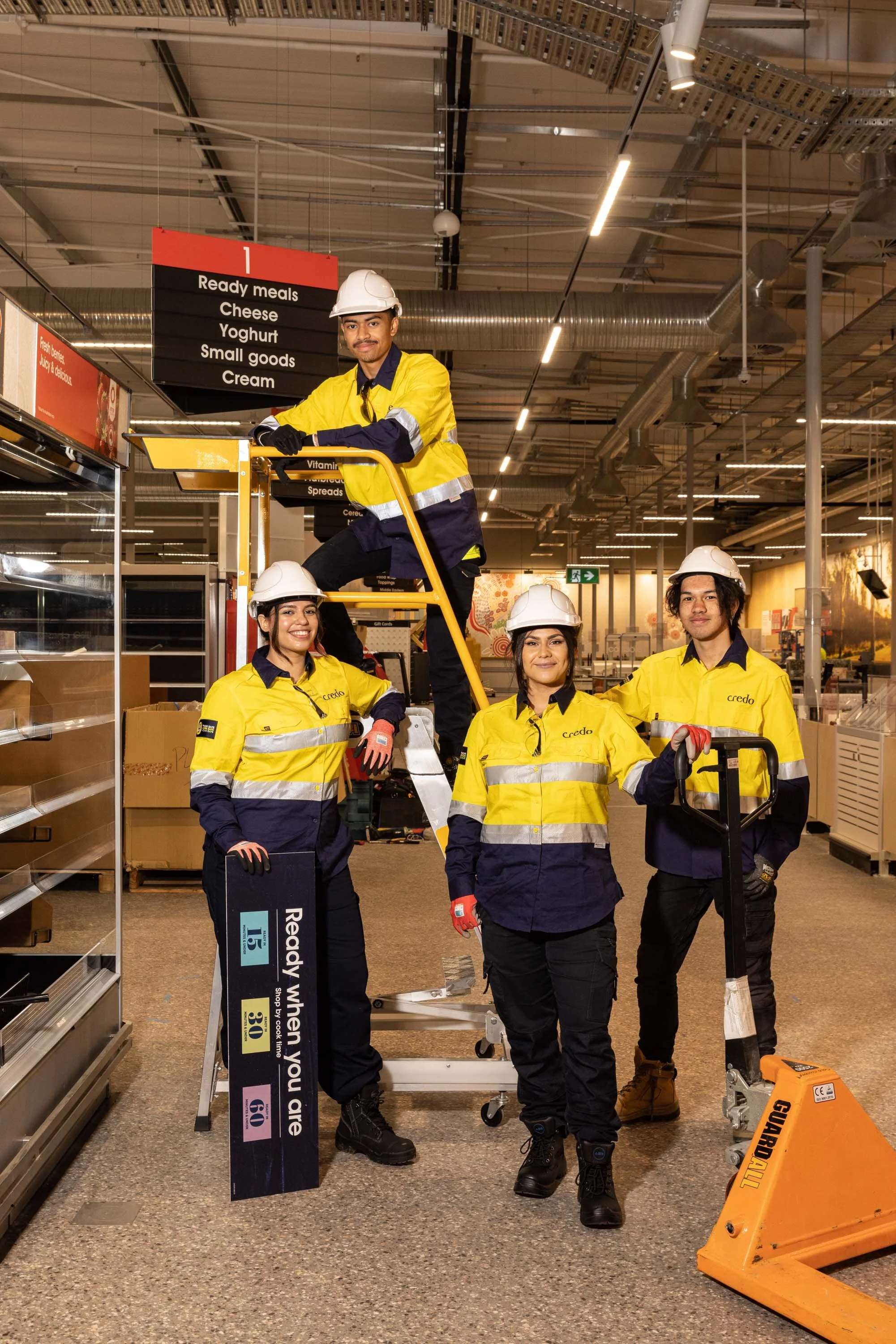 Group of five grocery store workers wearing yellow and navy uniforms with hard hats, posing and smiling inside a supermarket.