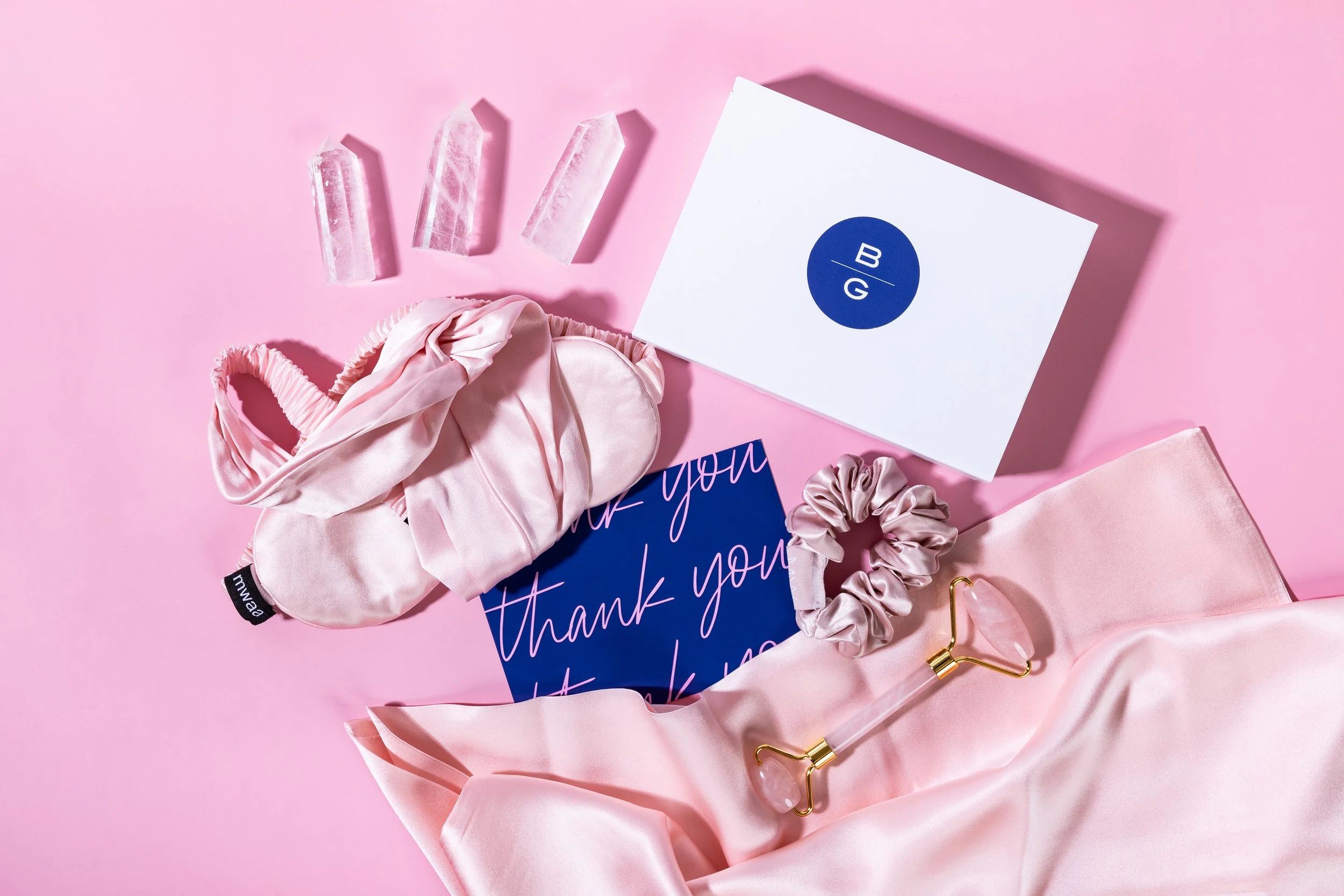 Pink silk eye mask, pink scrunchie, pink silk pillowcase, facial roller, thank you card, blue and white box with B and G logo, four pink crystals on pink background.