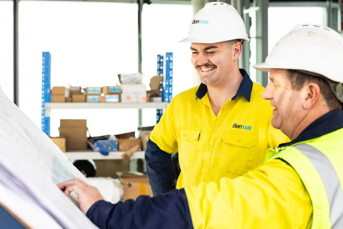 Two construction workers wearing yellow safety shirts and white hard hats at a construction site, examining blueprints and smiling.