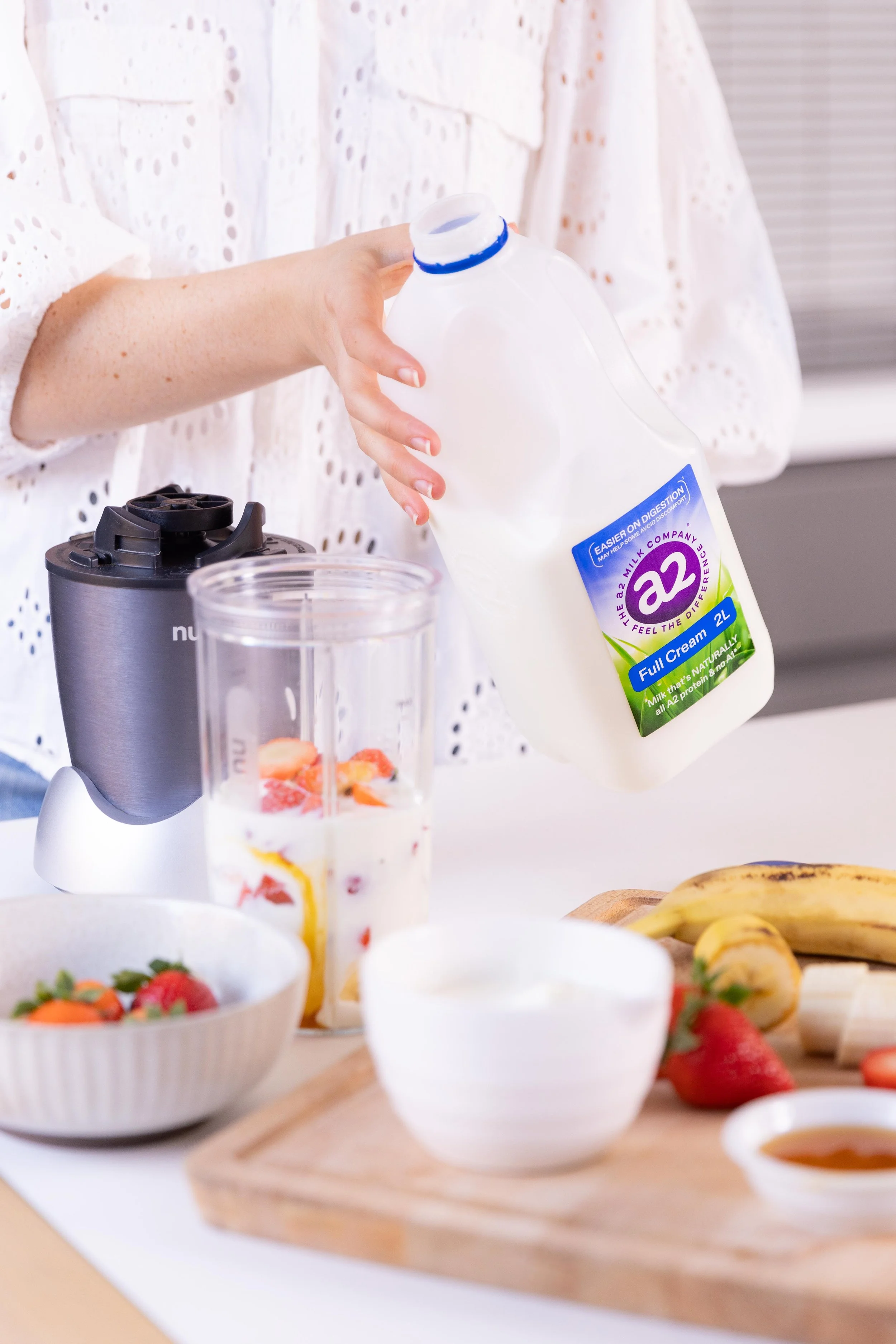 Person pouring milk into a blender on a kitchen counter with strawberries, bananas, and bowls of other fruits visible.
