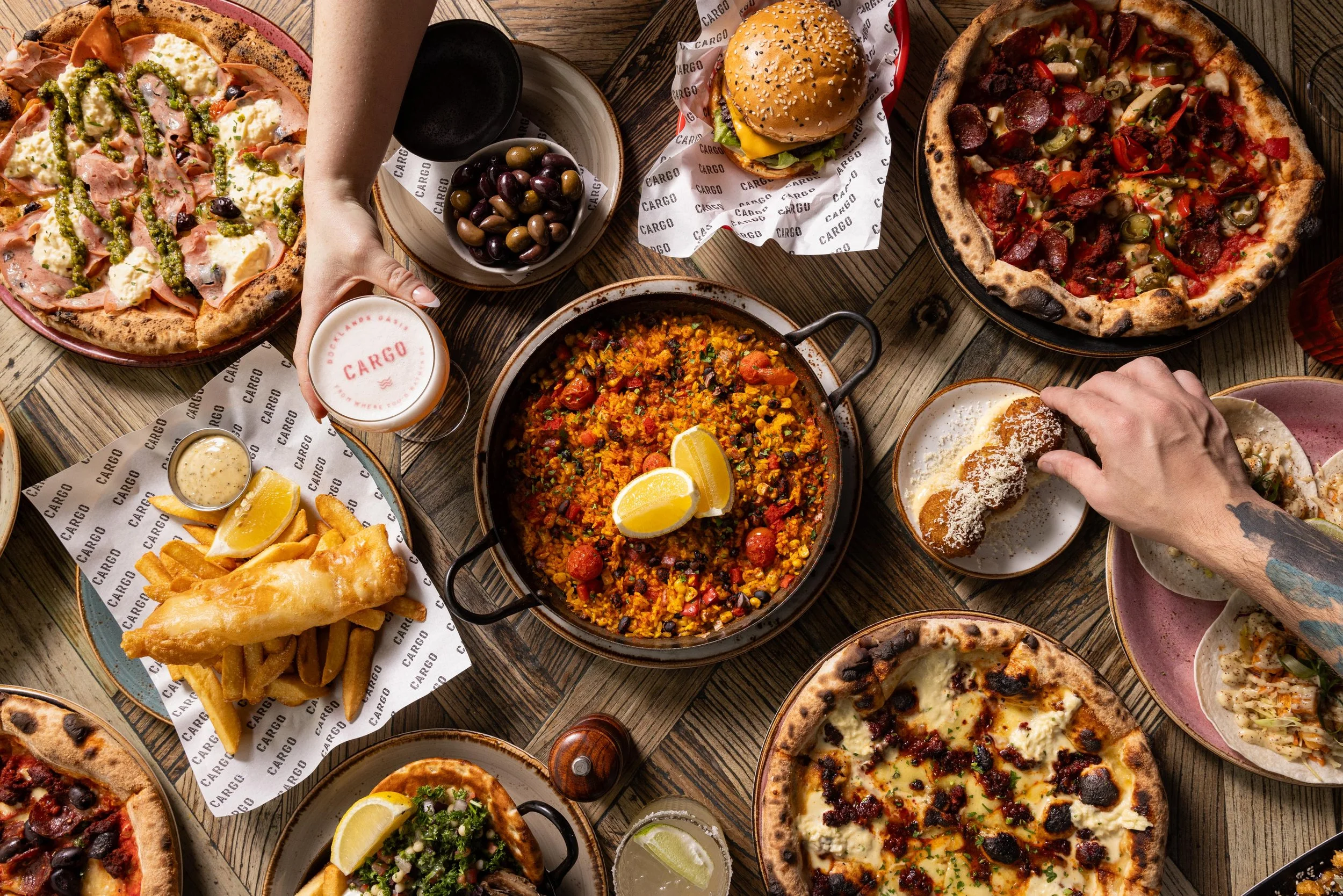 A large spread of food on a wooden table, including two pizzas, a burger, fried fish with fries, a rice dish with lemon wedges, a plate of tacos, and a bowl of olives, with drinks and condiments visible.