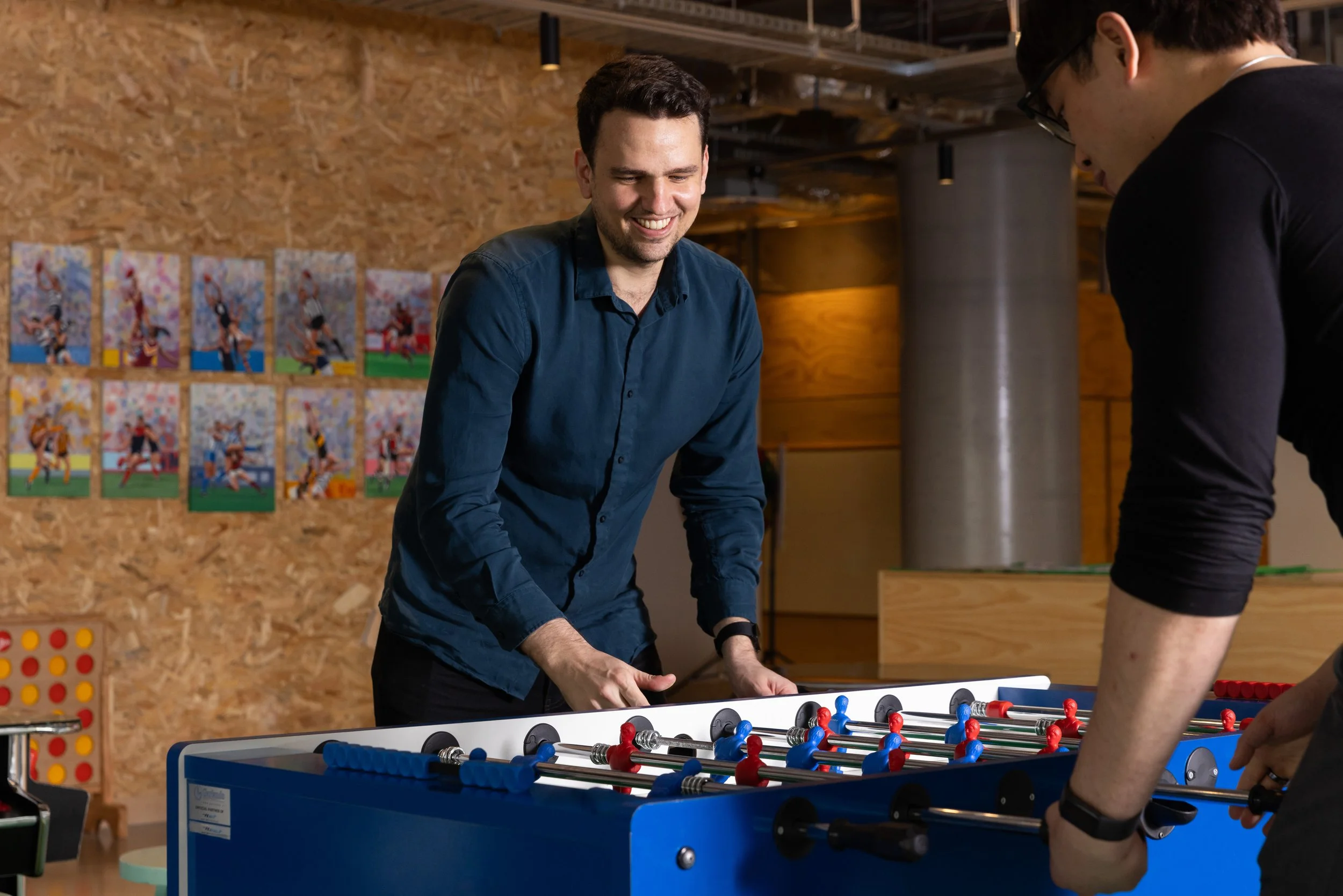 Two men playing foosball indoors, smiling. One wearing a blue shirt, the other a black shirt, with a wood-paneled wall and photos in the background.