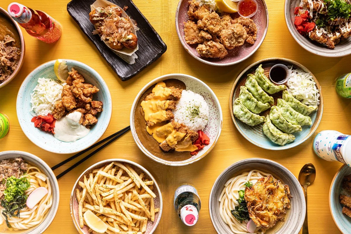 Assorted Japanese dishes on a yellow table, including curry rice, fried chicken, gyoza with dipping sauce, ramen, pasta, and various side dishes.