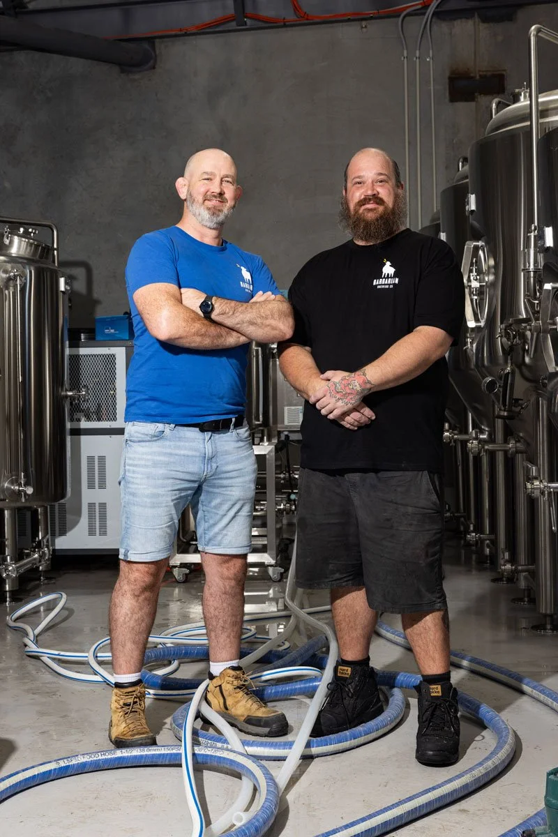Two men standing inside a brewery with large stainless steel brewing tanks and various pipes and hoses on the floor.