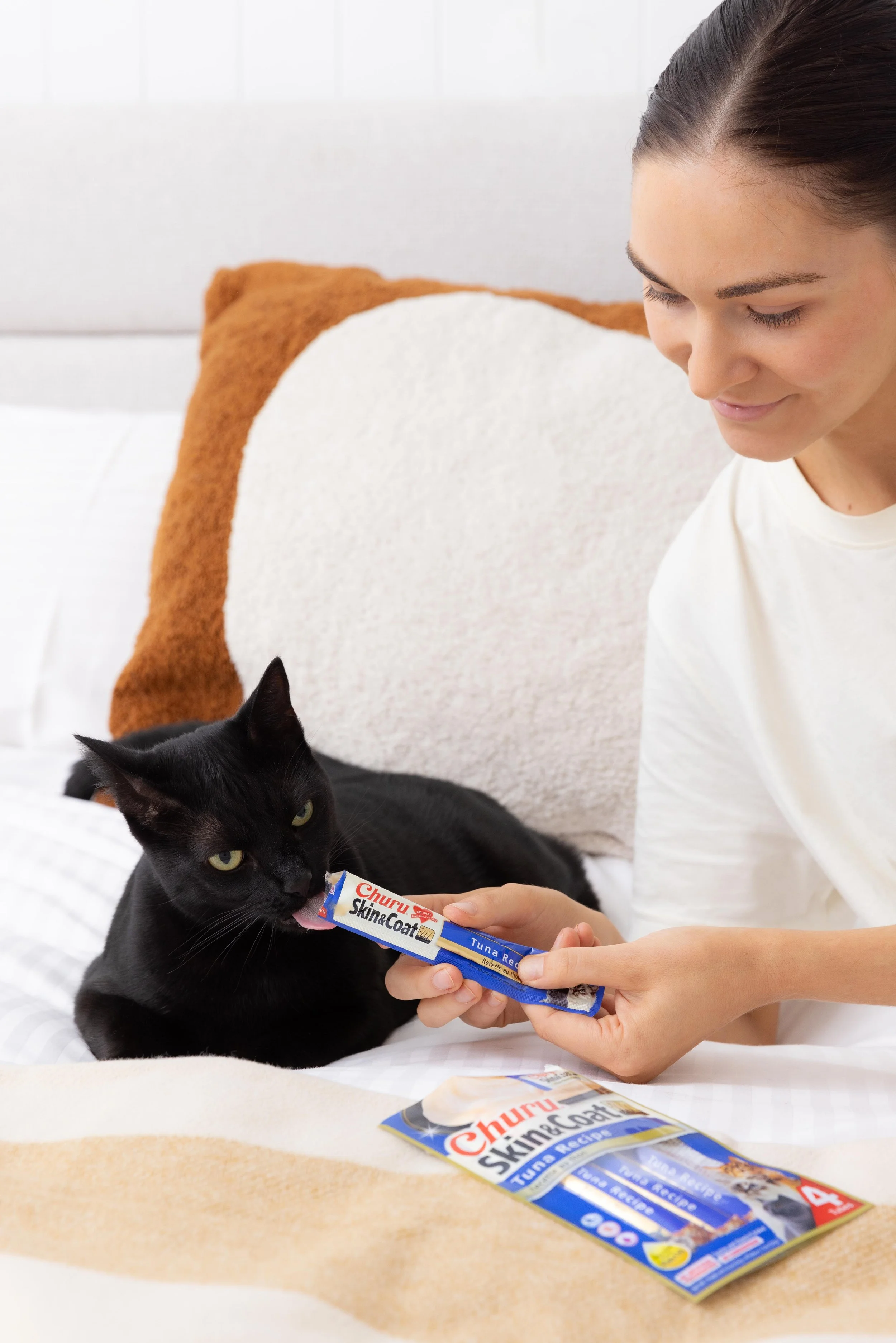A woman feeds a black cat a tube of tuna-flavored cat food on a bed with pillows.