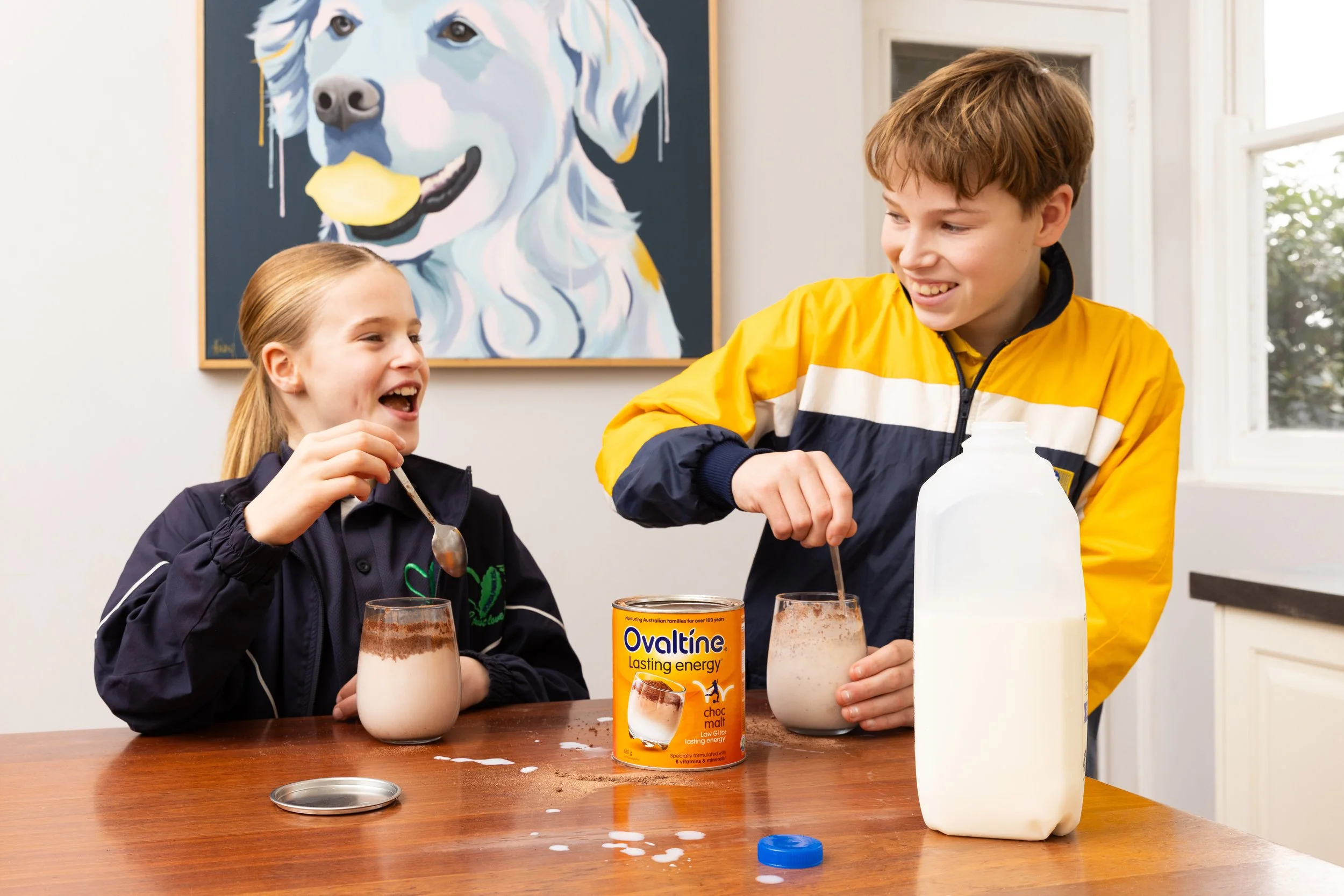 A boy and girl mixing milk and chocolate powder for a chocolate milk drink at a wooden table, with a large cartoon dog painting in the background.