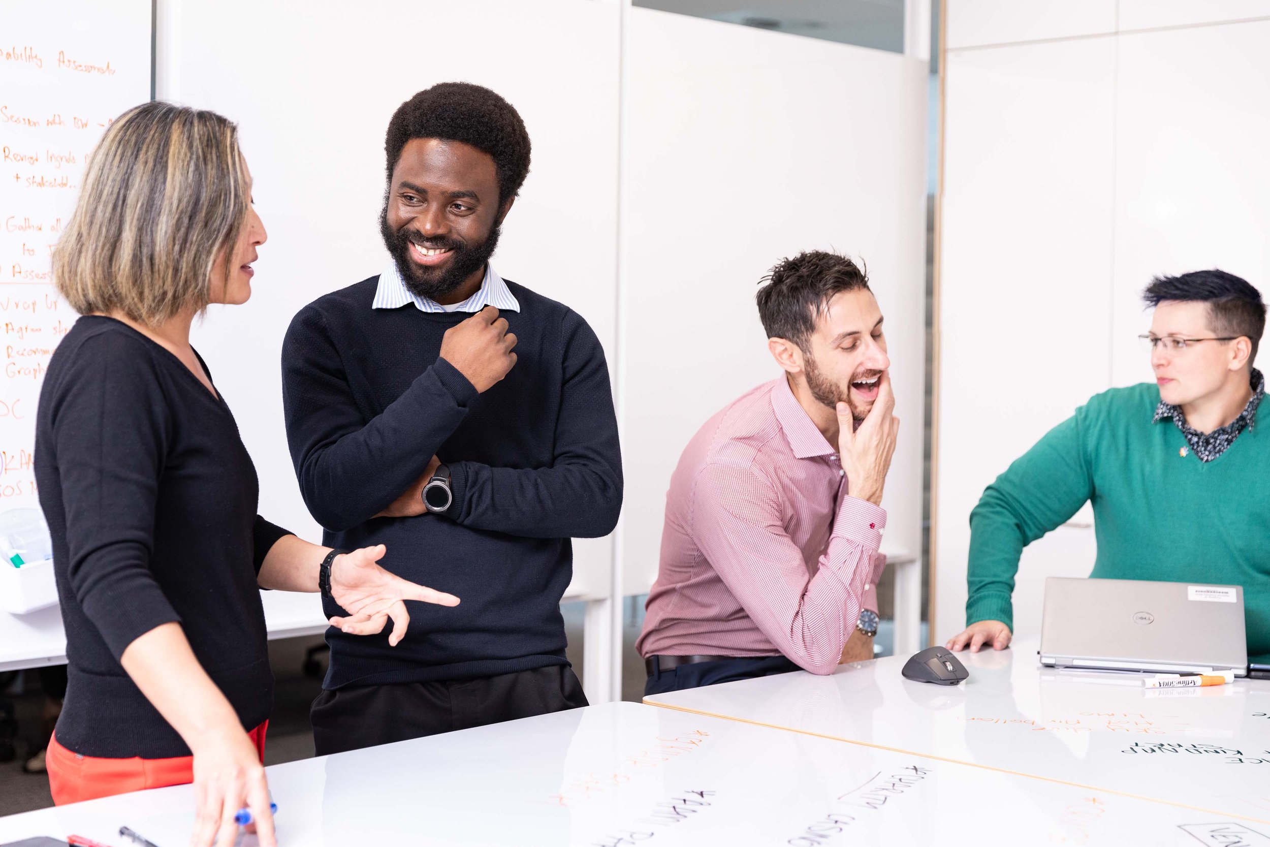 Four people in a meeting room, engaging in conversation and laughter. One woman is speaking, and three men are reacting with smiles, one touching his chin, and one leaning on a laptop.