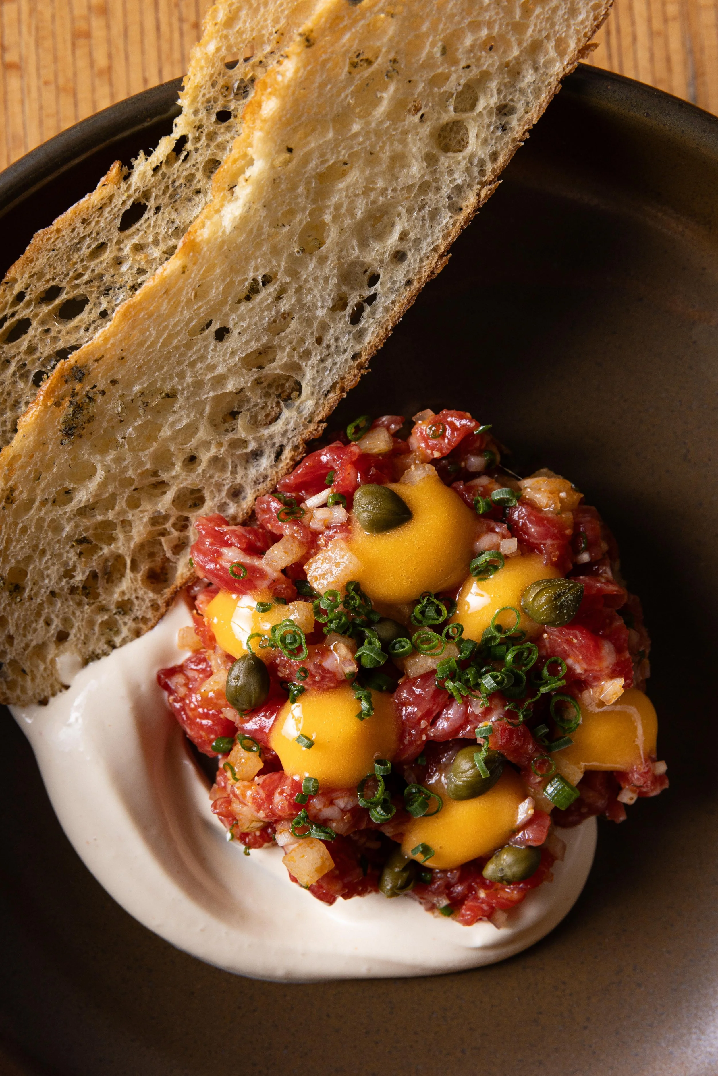 A dish of steak tartare topped with yellow egg yolks, capers, chopped chives, served with a dollop of cream and two slices of toasted bread on a brown plate.