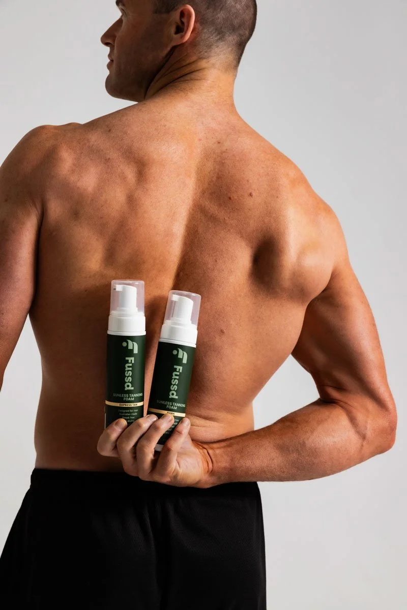 Man with tan skin holding two cans of Fuss facial tanning foam behind his back, shirtless, against a light background.