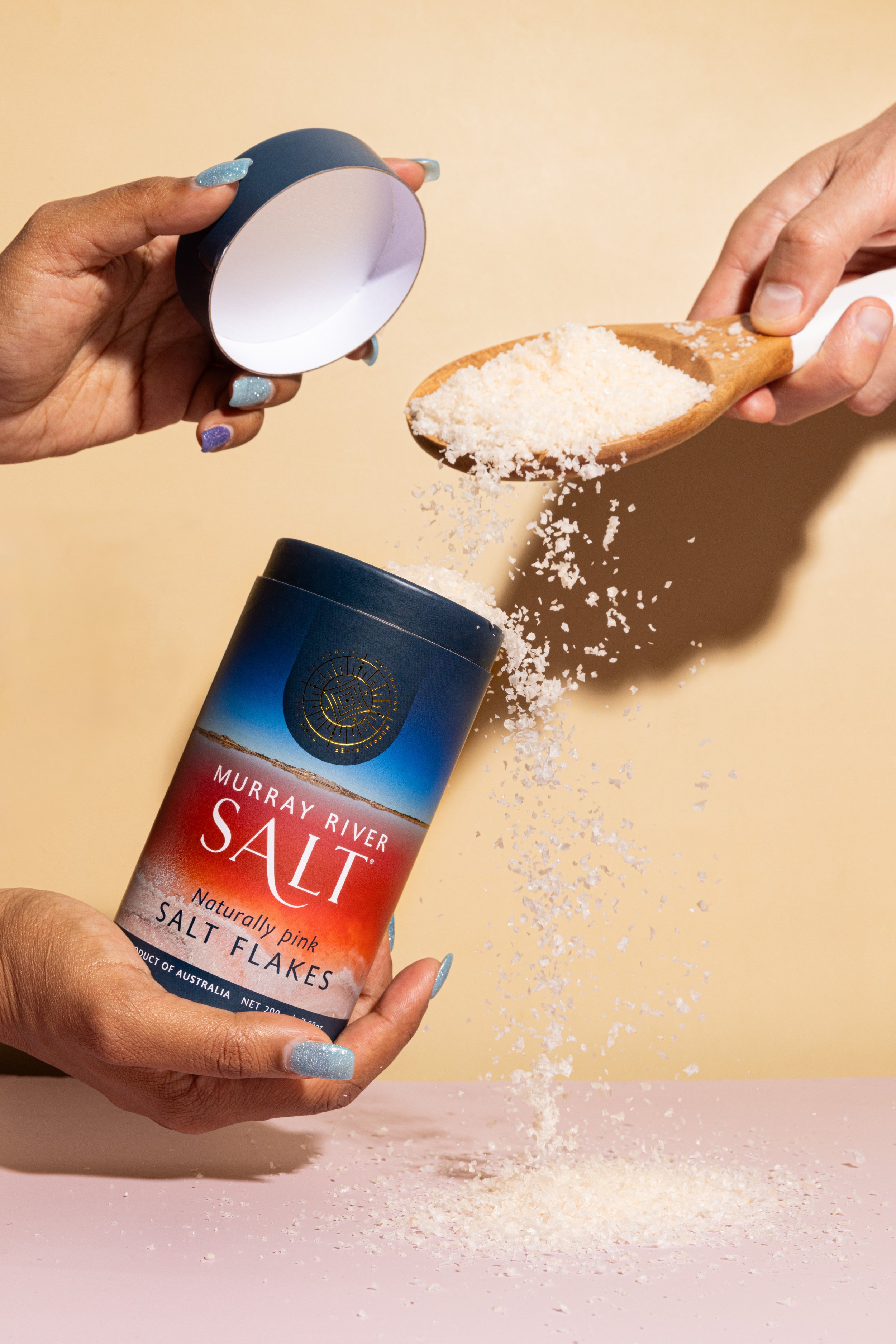 Hands pouring pink salt flakes from a wooden spoon into a container labeled 'Murray River Salt' with a blue and red sunset background.