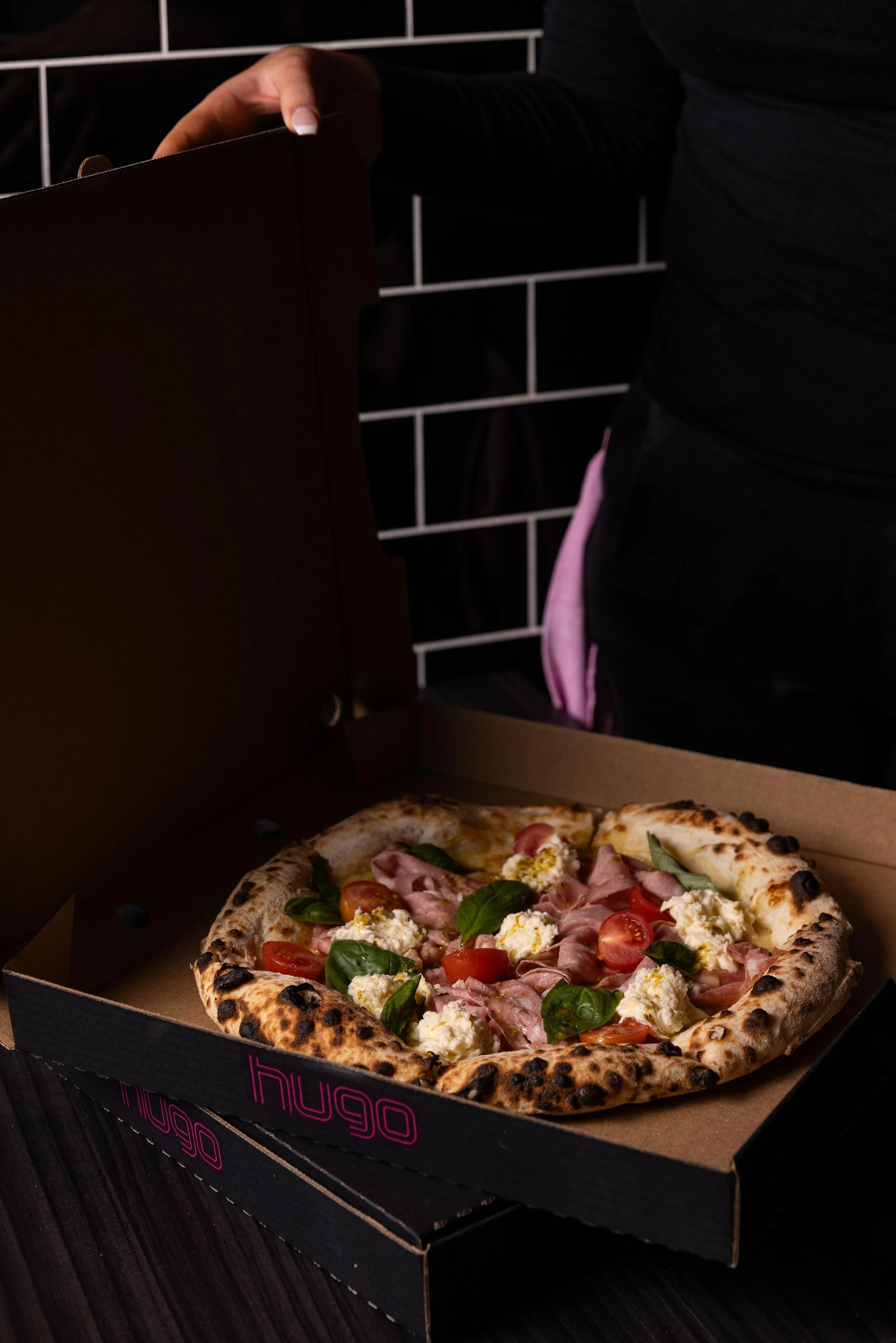 A person opening a black pizza box with a freshly baked pizza containing cherry tomatoes, fresh basil, ricotta cheese, ham, and melted cheese.