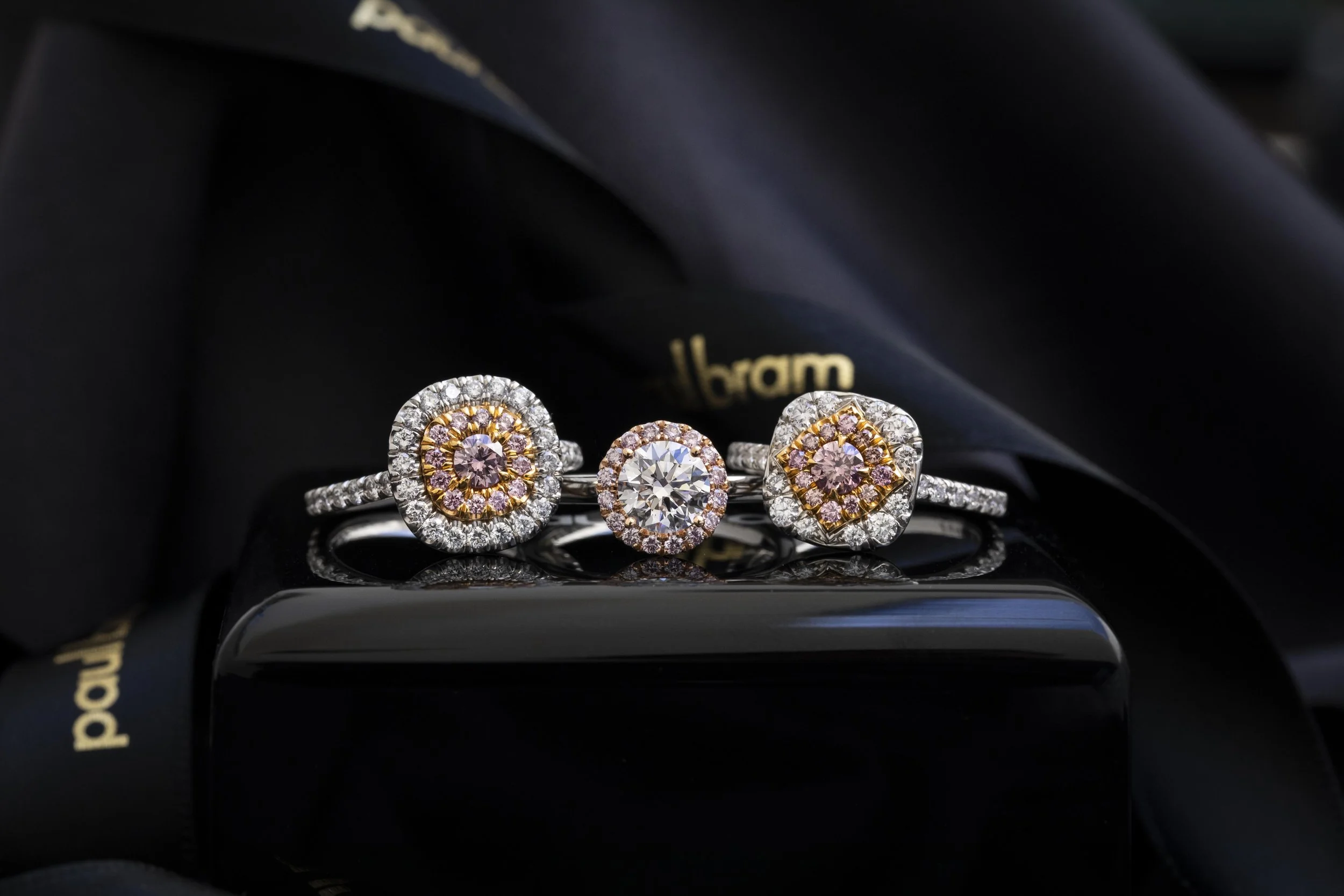 Three diamond rings with pink and white stones, placed on a black reflective surface, with gold and black packaging in the background.