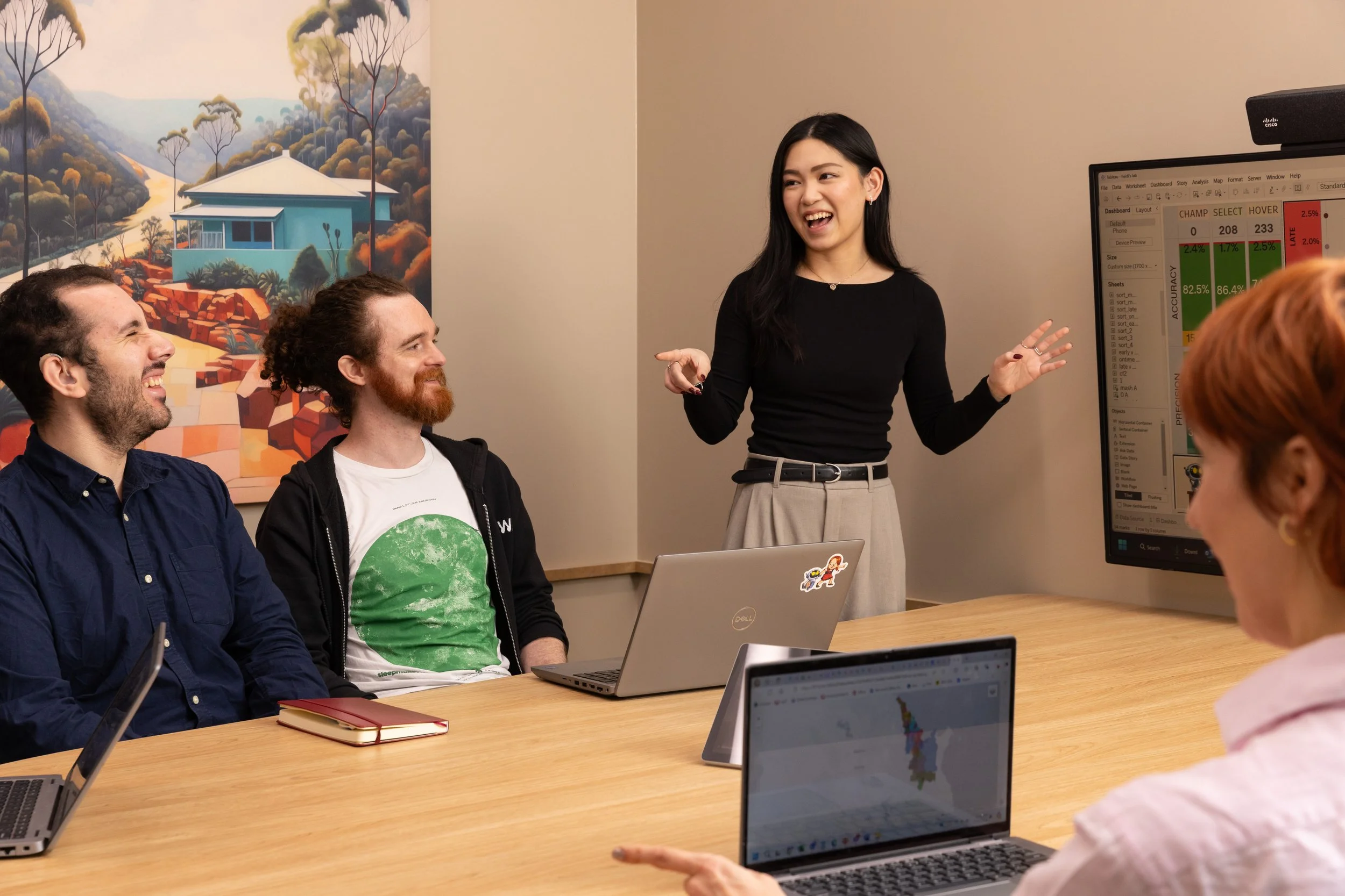 A woman presenting to a group of people in a meeting room, with a large monitor displaying data, and other laptops on the table.