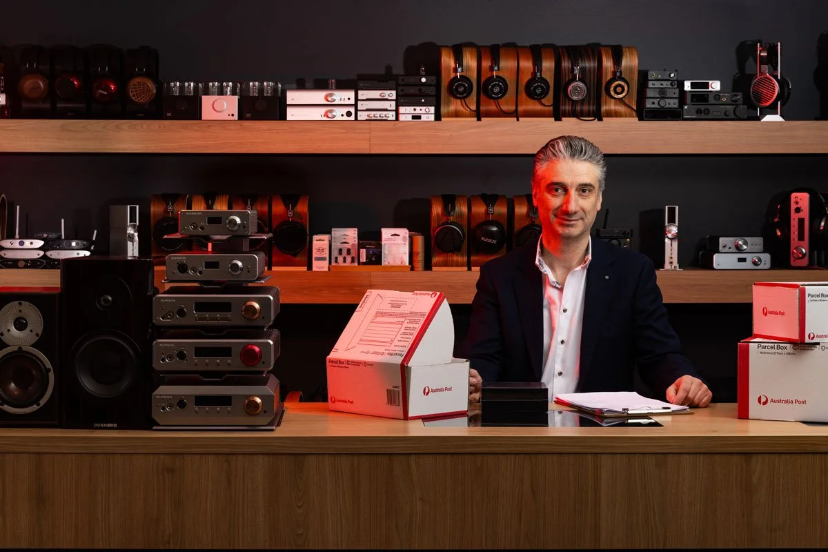A man sitting behind a wooden counter surrounded by electronic devices, including speakers, audio equipment, headphones, and postal boxes labeled Australia Post.