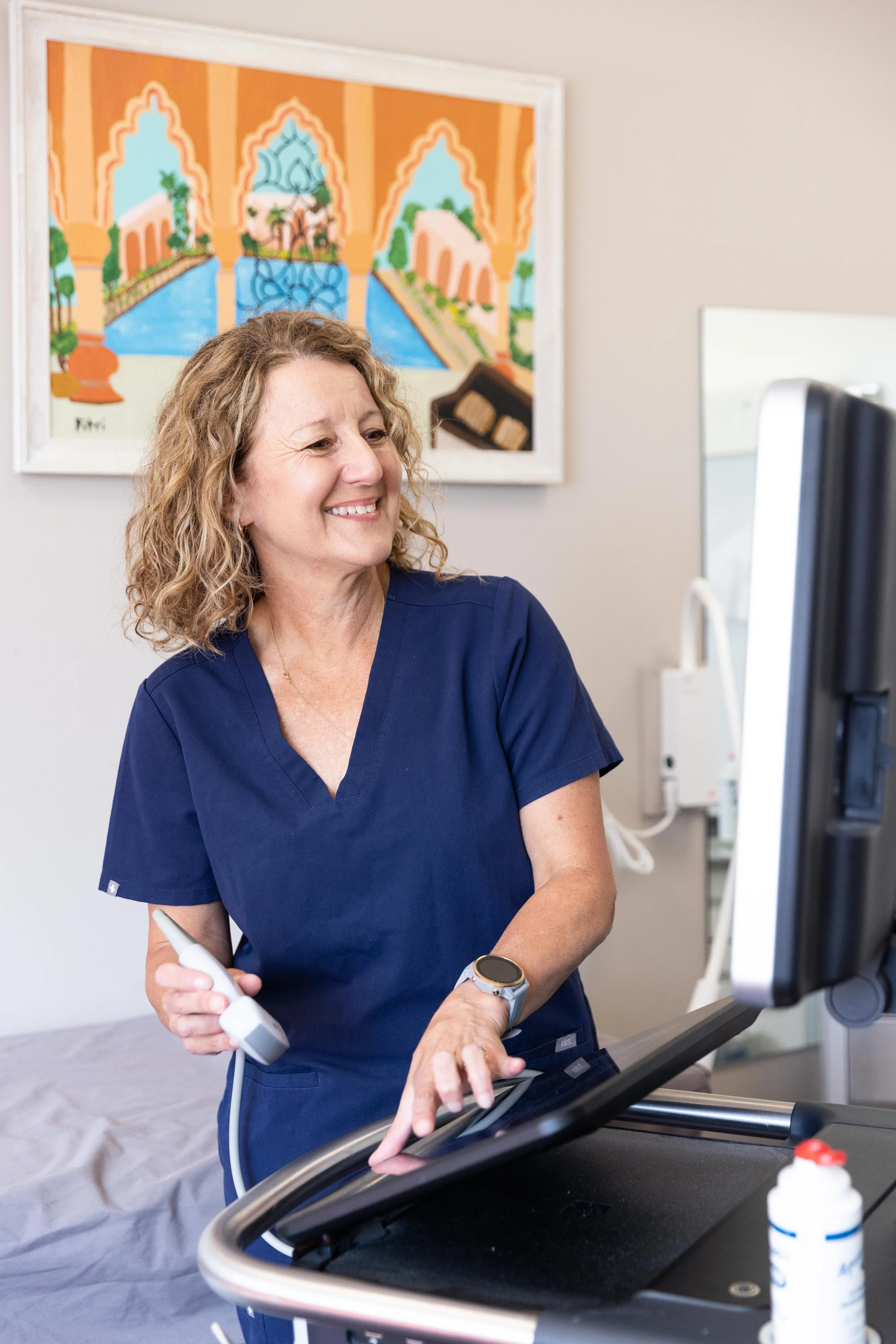 A healthcare professional, wearing navy scrubs, standing next to an ultrasound machine, smiling and looking at the monitor in a medical setting.