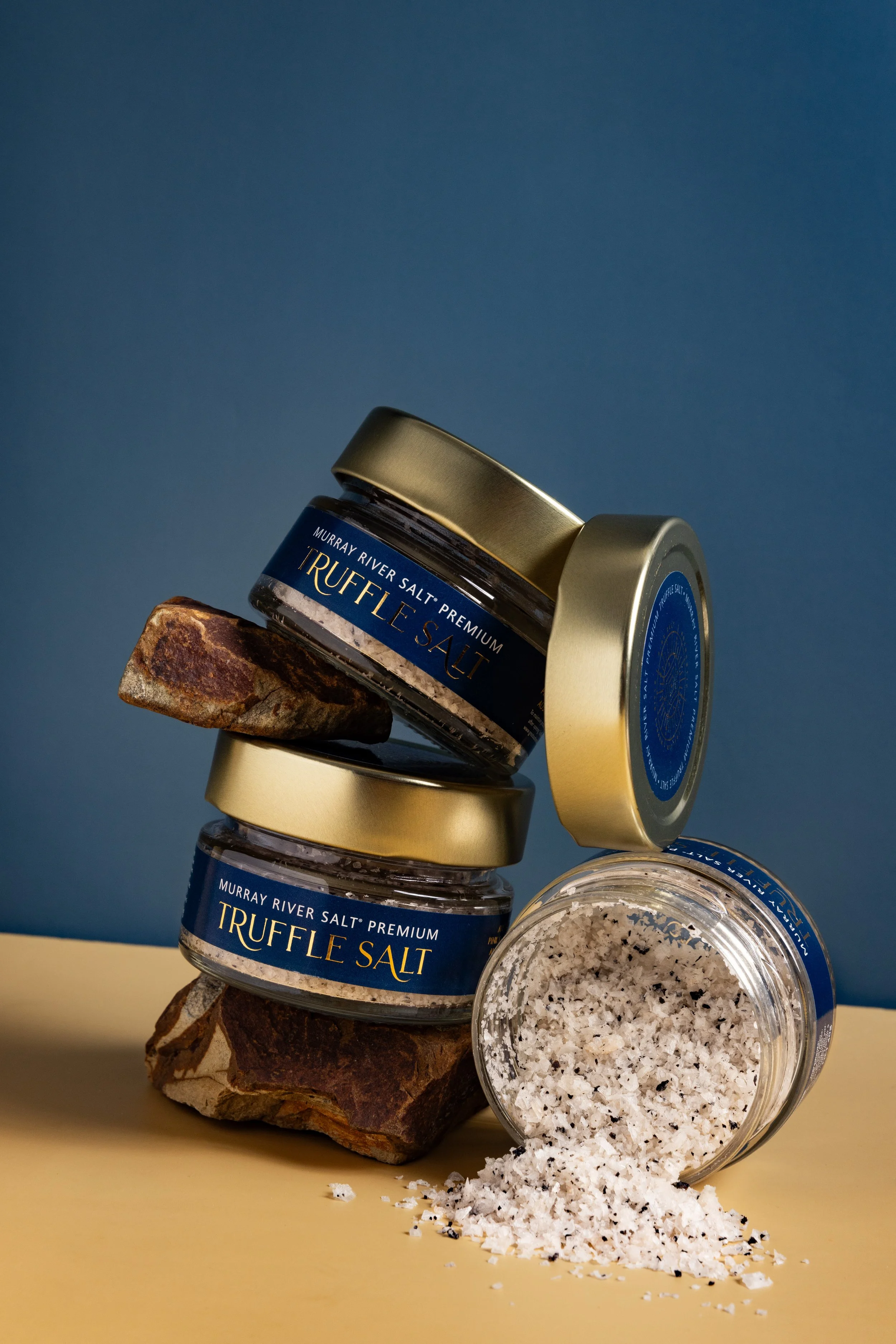 Two small jars of Murray River Salt Truffle Salt, one tilted with black salt and flakes spilling onto a yellow surface, supported by rocks, with a blue and yellow background.