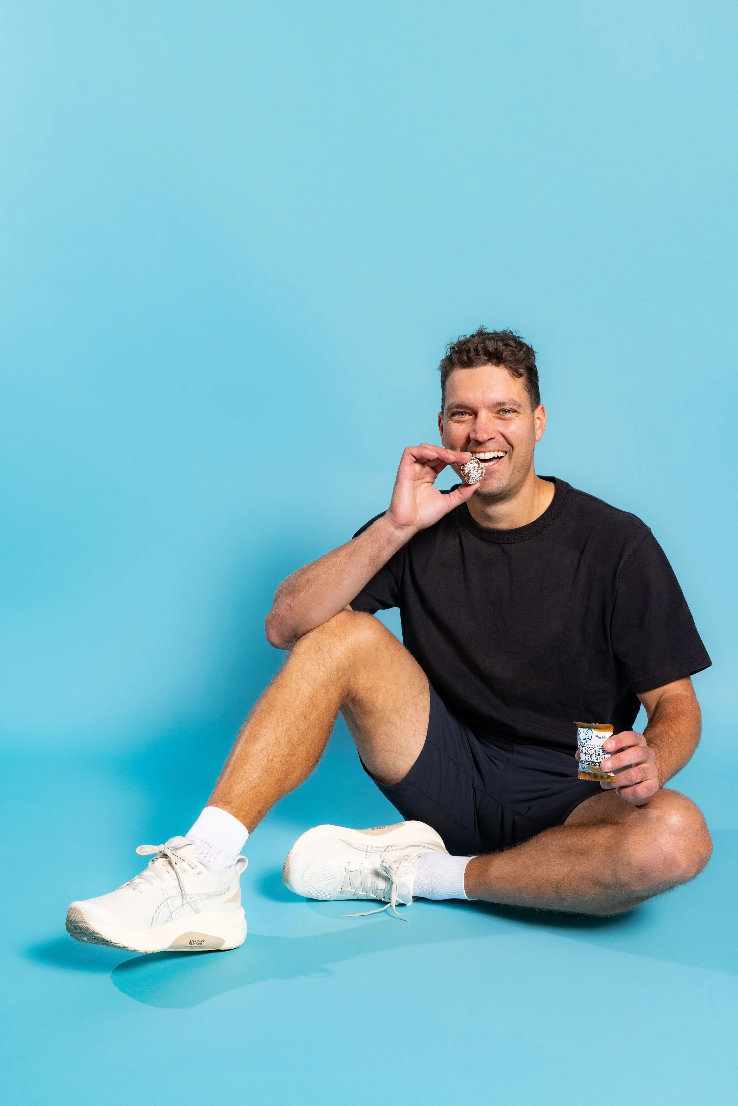 A smiling man sitting on a blue floor and background, eating a chocolate snack and holding a container of snacks, dressed in a black t-shirt, black shorts, white sneakers, and white socks.