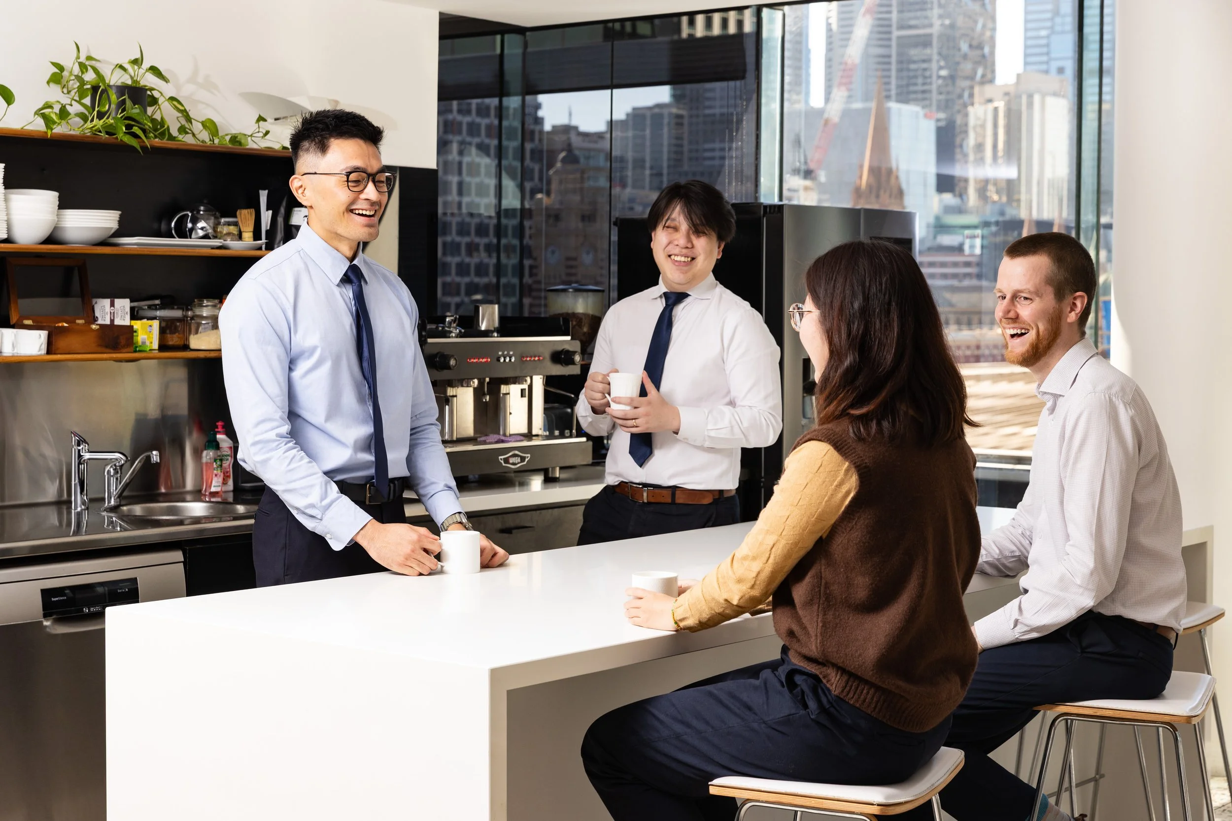 Four coworkers laughing and chatting in an office break room with large windows, a cityscape view, and a coffee station.