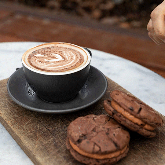 A black cup of latte with latte art on top, placed on a saucer on a wooden board, with two chocolate cookies nearby on a marble surface.