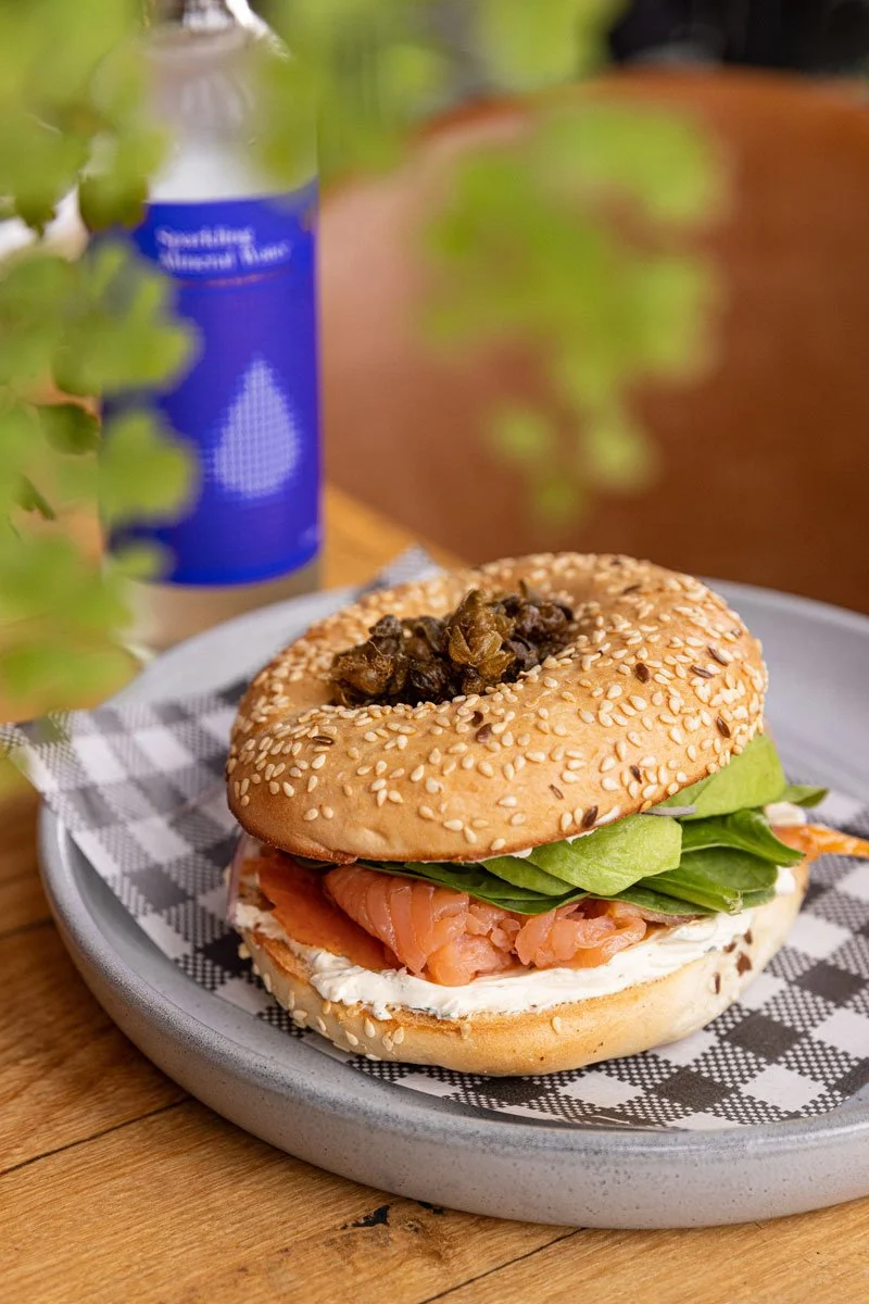A bagel sandwich with smoked salmon, cream cheese, lettuce, and a topping of chopped olives on a sesame seed bagel, placed on a checkered napkin on a gray plate.