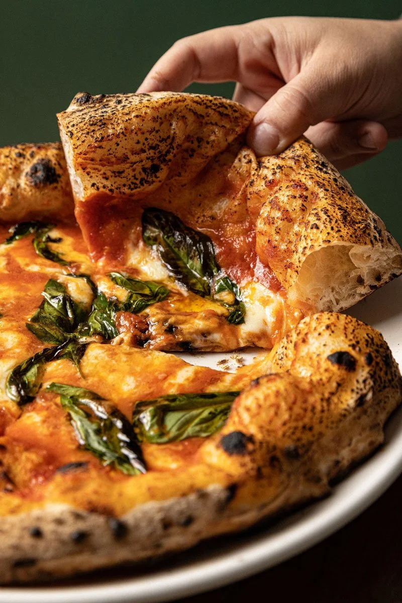 Close-up of a hand lifting a slice of Margherita pizza with melted mozzarella cheese, tomato sauce, basil leaves, and a charred crust.