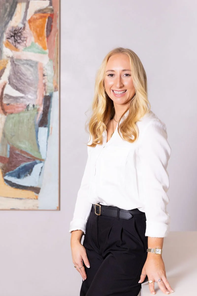 A young woman with long blonde hair wearing a white blouse and black pants, smiling, standing indoors next to an abstract painting.