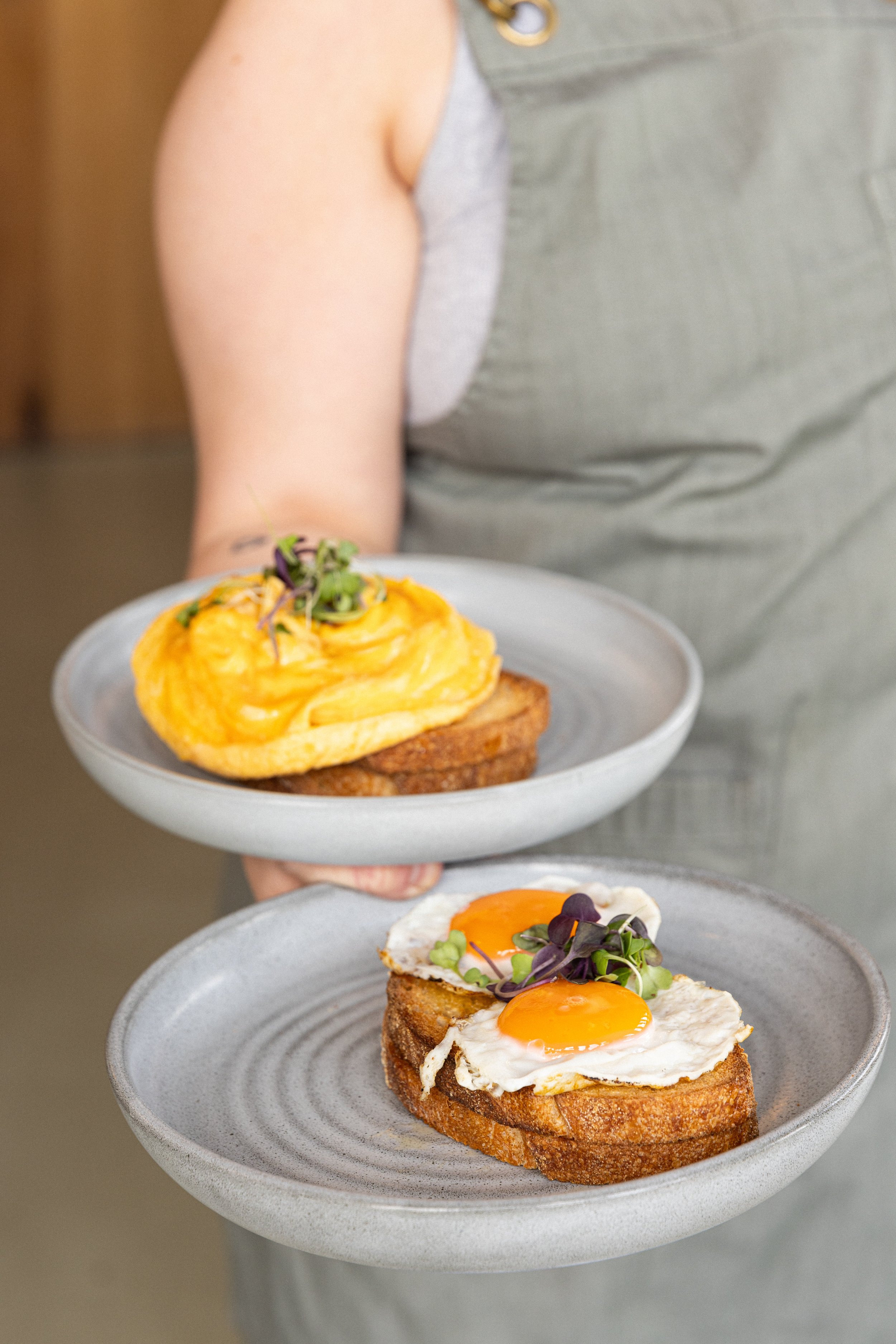 Person holding two plates of breakfast dishes, one with eggs Benedict topped with microgreens and the other with scrambled eggs on toast with microgreens.