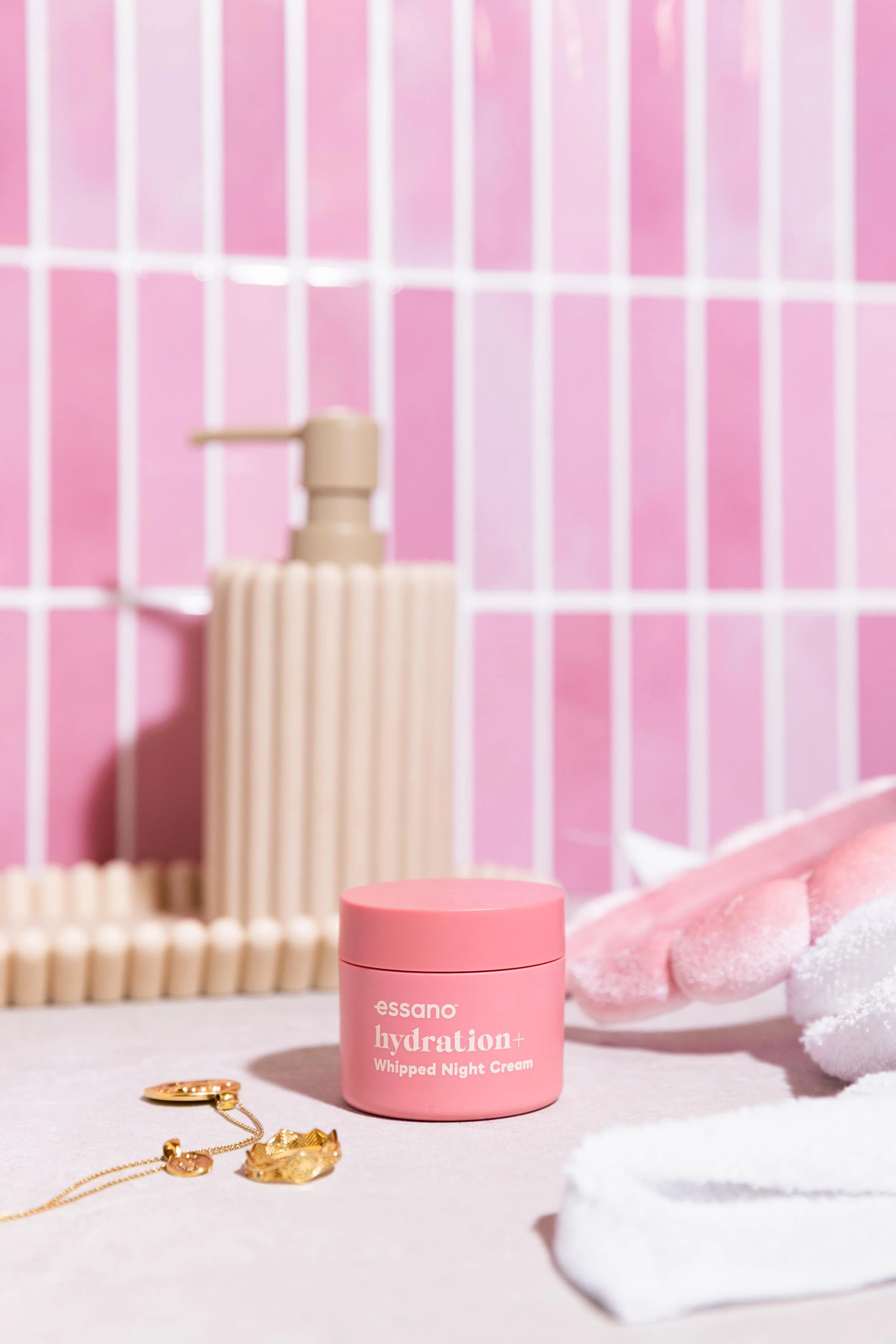 A pink jar of Essano Hydration+ Whipped Night Cream on a white surface with pink and white towels and makeup brushes in the background