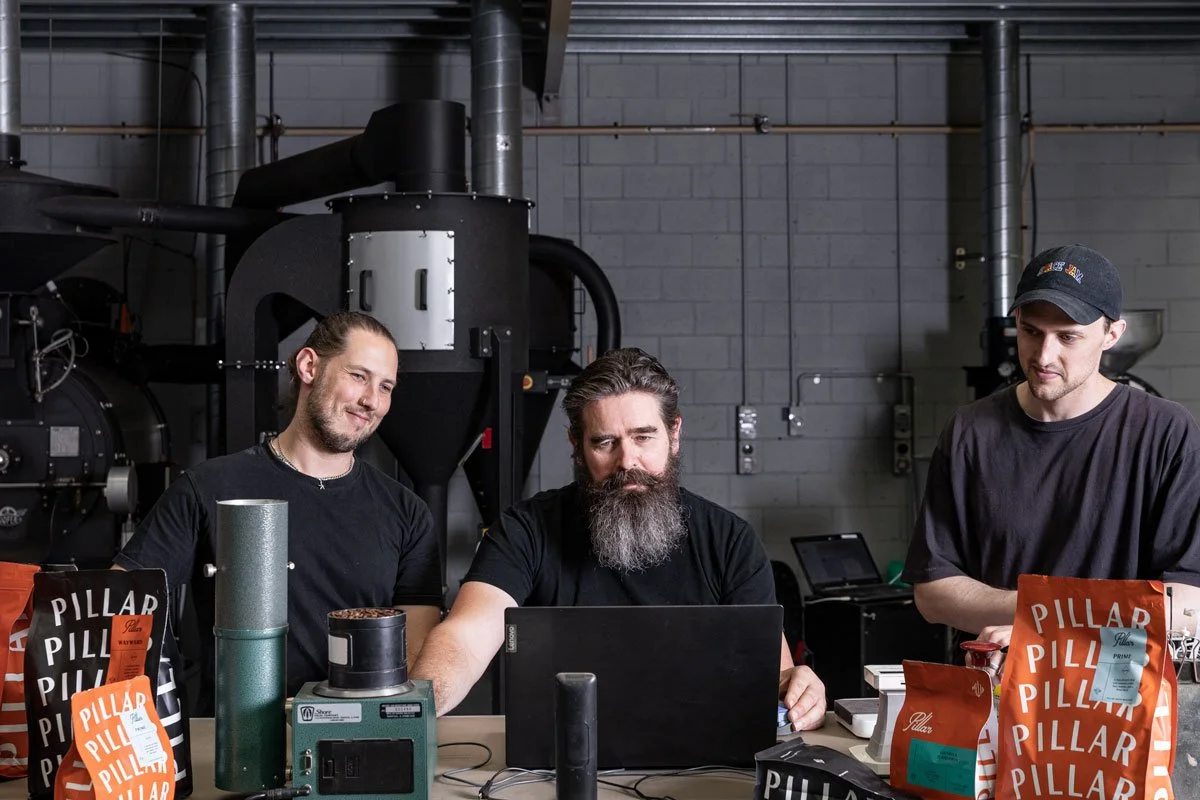 Three men sitting at a table with coffee and snack bags, working on a laptop in an industrial setting with machinery and pipes in the background.