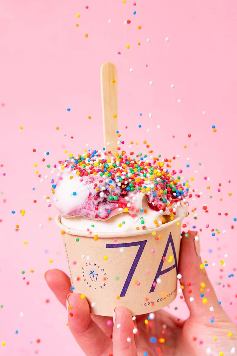 A cup of ice cream with colorful sprinkles on top, held in a person's hand against a pink background with sprinkles falling around.