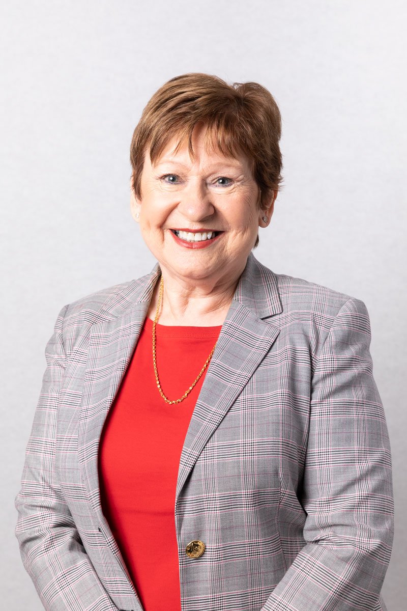 A smiling older woman with short brown hair, wearing a red top, a plaid blazer, a gold necklace, and small earrings, standing in front of a plain light background.