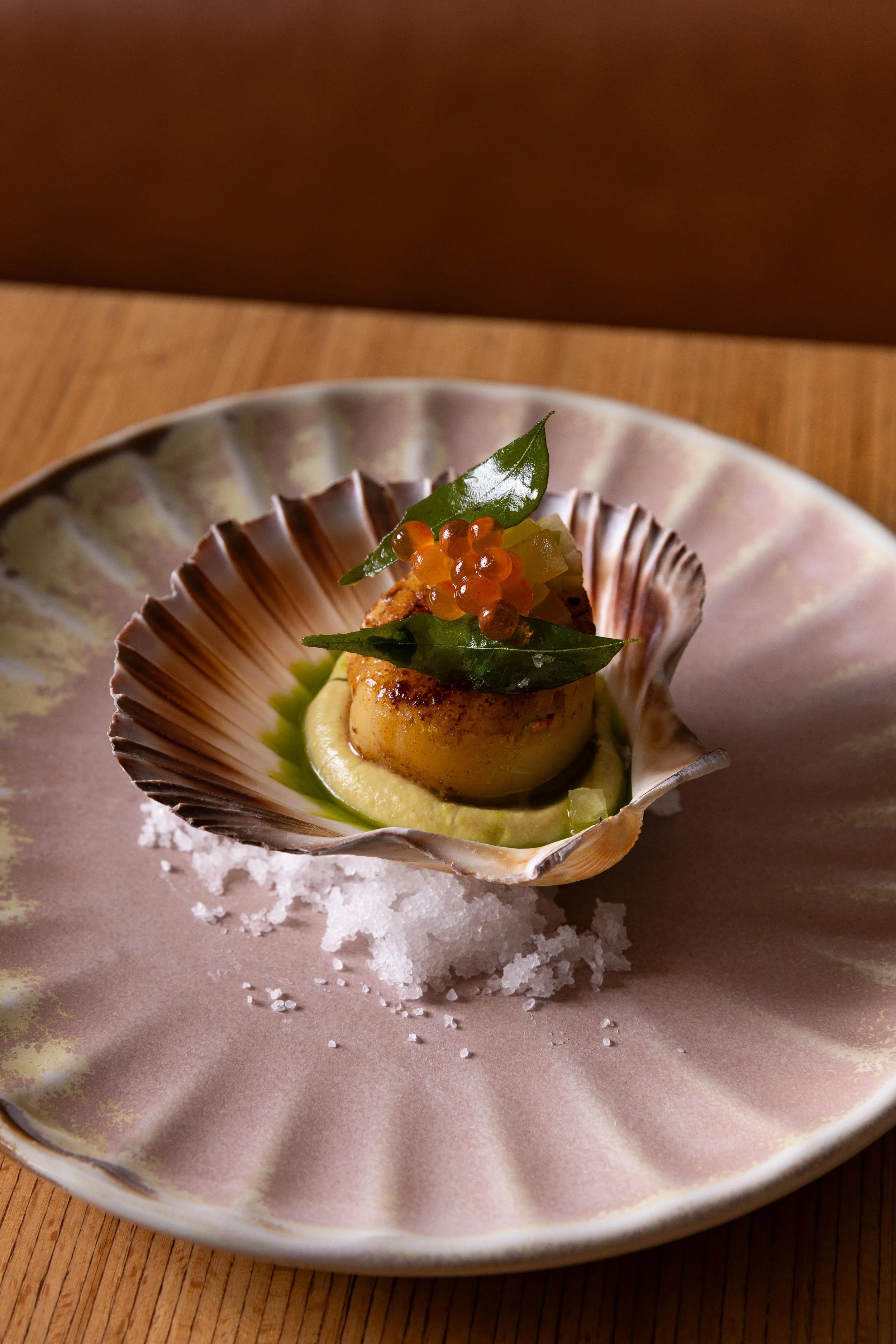 Seared scallop served inside a scallop shell on a ceramic plate with salt flakes and green sauce, garnished with small orange fish roe, green leaves, and diced vegetables.