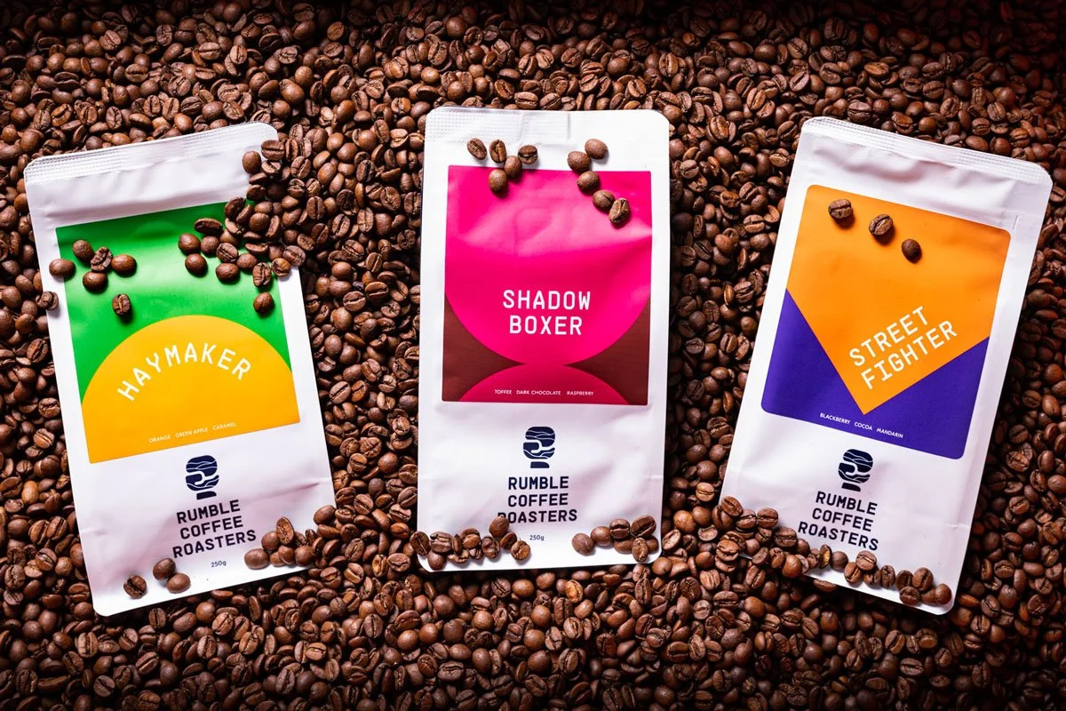 Three bags of Rumble Coffee Roasters coffee beans lie on a bed of coffee beans. The bag on the left is labeled 'Haymaker' with a green and yellow design, the middle bag is labeled 'Shadow Boxer' with a pink and brown design, and the right bag is labe