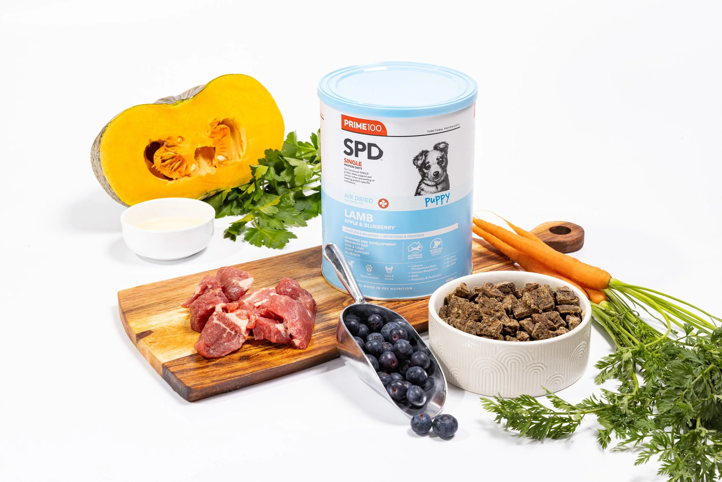 Dog food ingredients including canned dog food, raw meat, blueberries, carrots, pumpkin, herbs, and a small bowl of yogurt on a white background.