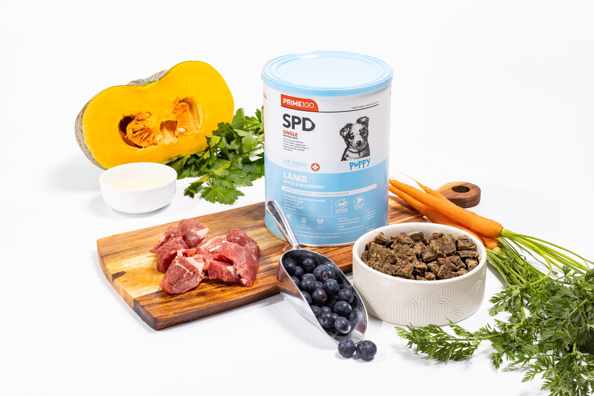Dog food ingredients including canned dog food, raw meat, blueberries, carrots, pumpkin, herbs, and a small bowl of yogurt on a white background.