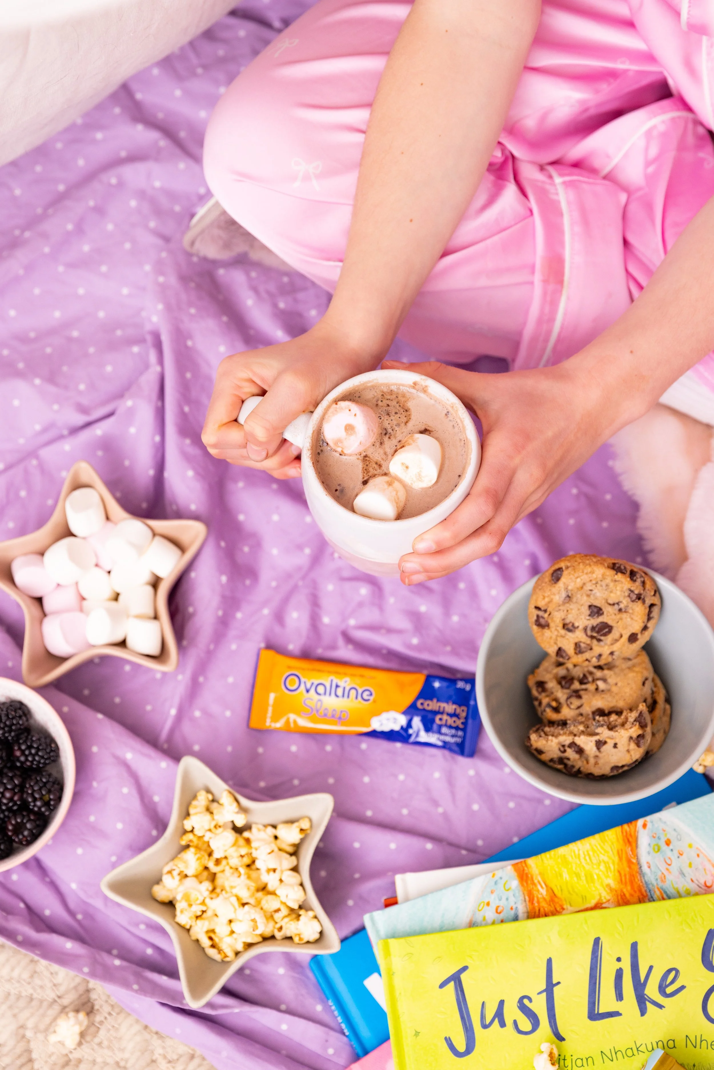 A person holding a mug of hot chocolate with marshmallows, surrounded by bowls of popcorn, blackberries, and marshmallows, on a purple polka-dot blanket with children's books nearby.