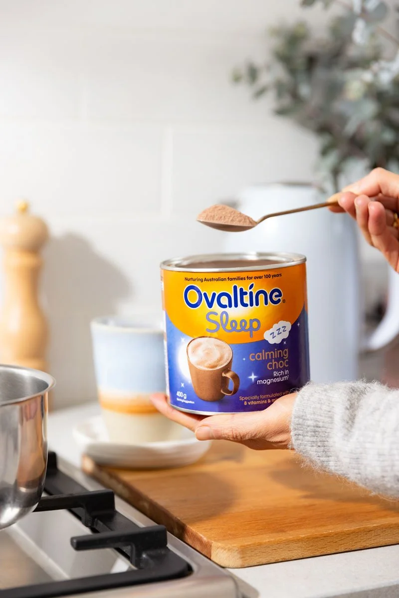 A person is holding a can of Ovaltine Sleep calming hot chocolate mix over a wooden cutting board in a kitchen. The person is about to add powder from a spoon into the can.