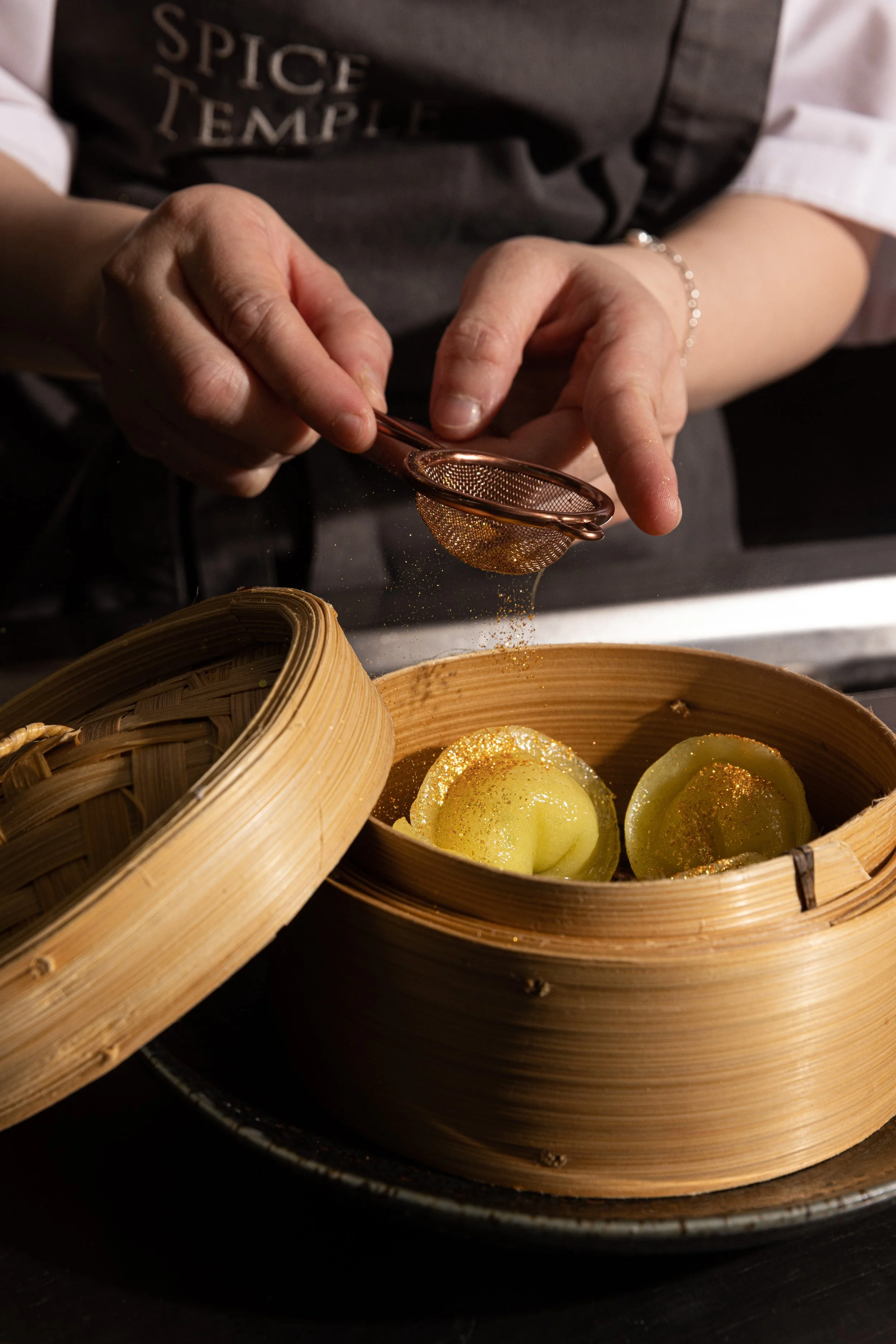 A person sprinkling a spice or seasoning over melon balls in a bamboo steamer basket.